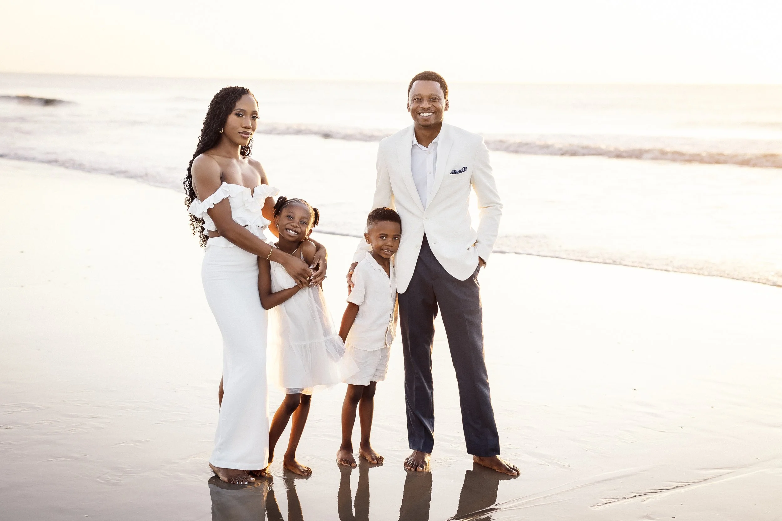 A family of five standing on the beach during sunset, dressed in white and light-colored clothing, smiling and posing for a photo.
