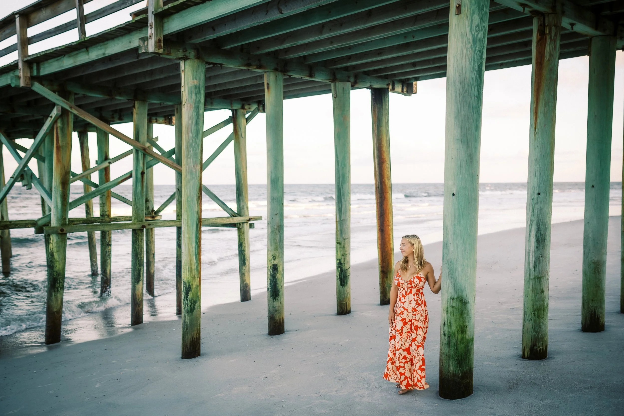 A woman in a floral orange dress standing under a pier on the beach, near the ocean.