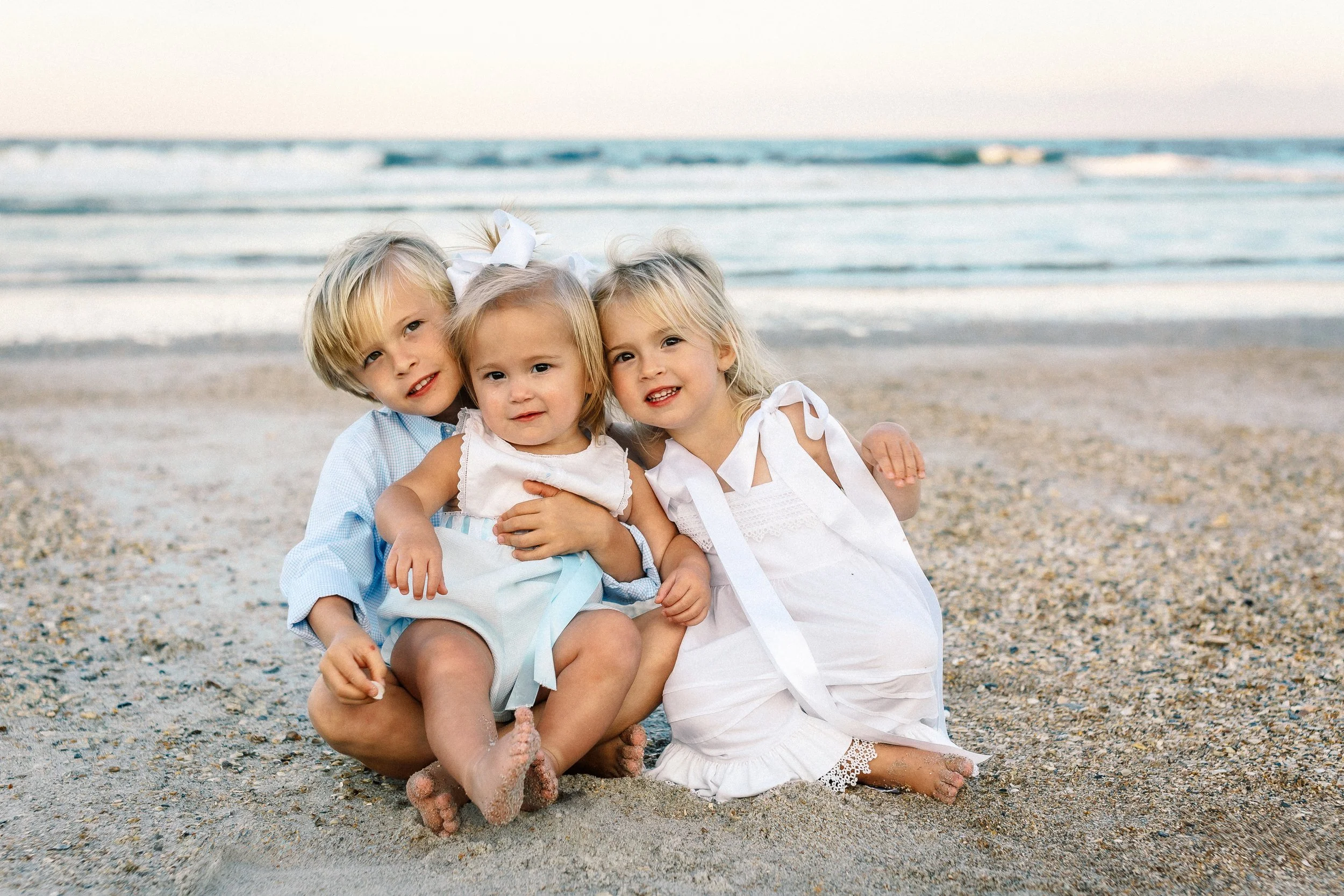 Four young children sitting on a sandy beach, smiling at the camera, with ocean waves in the background.Photographer Amelia Island, Jacksonville, St Augustine, St Simons Island, Savannah photographer