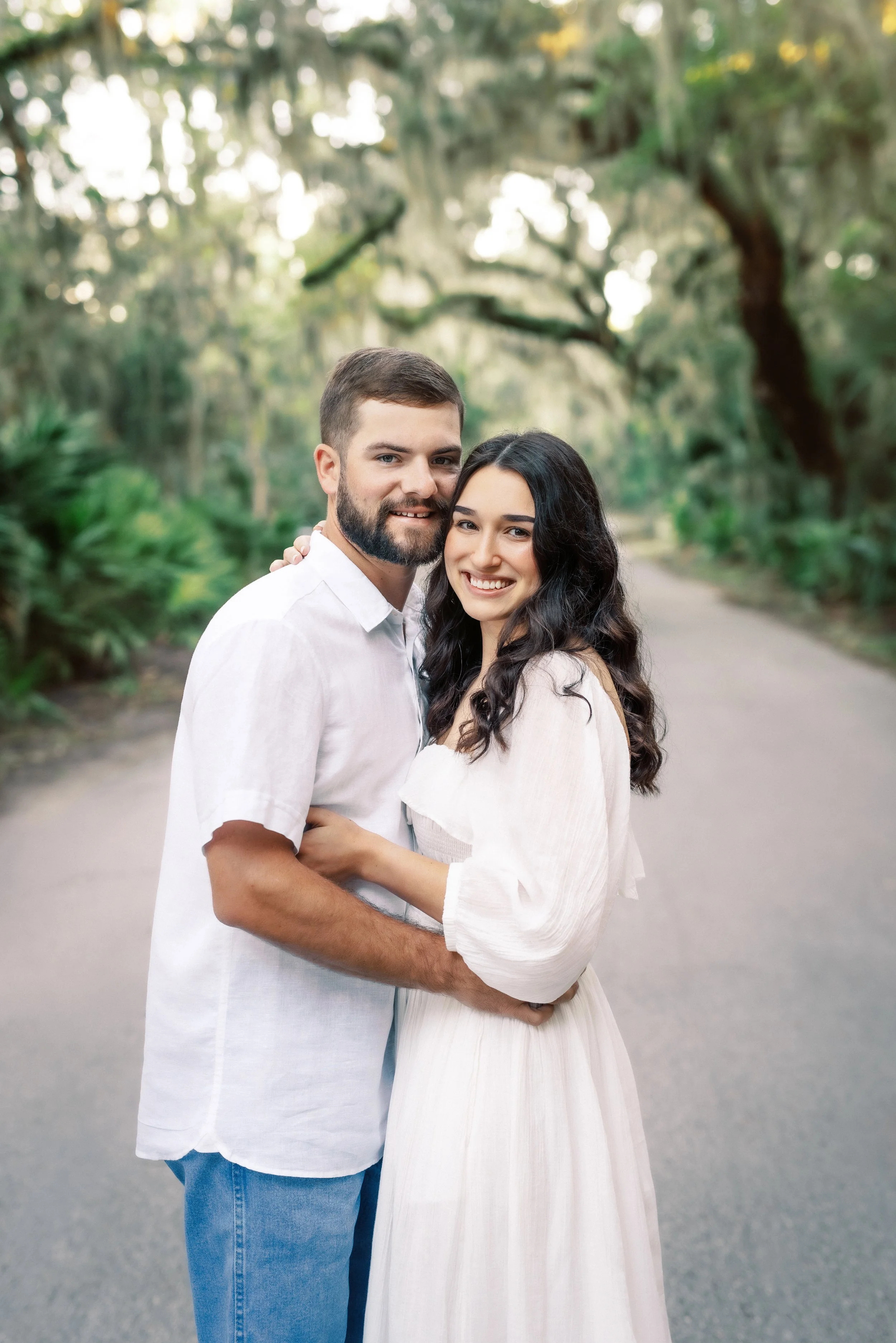A smiling couple standing close together outdoors on a pathway, surrounded by green trees and foliage.