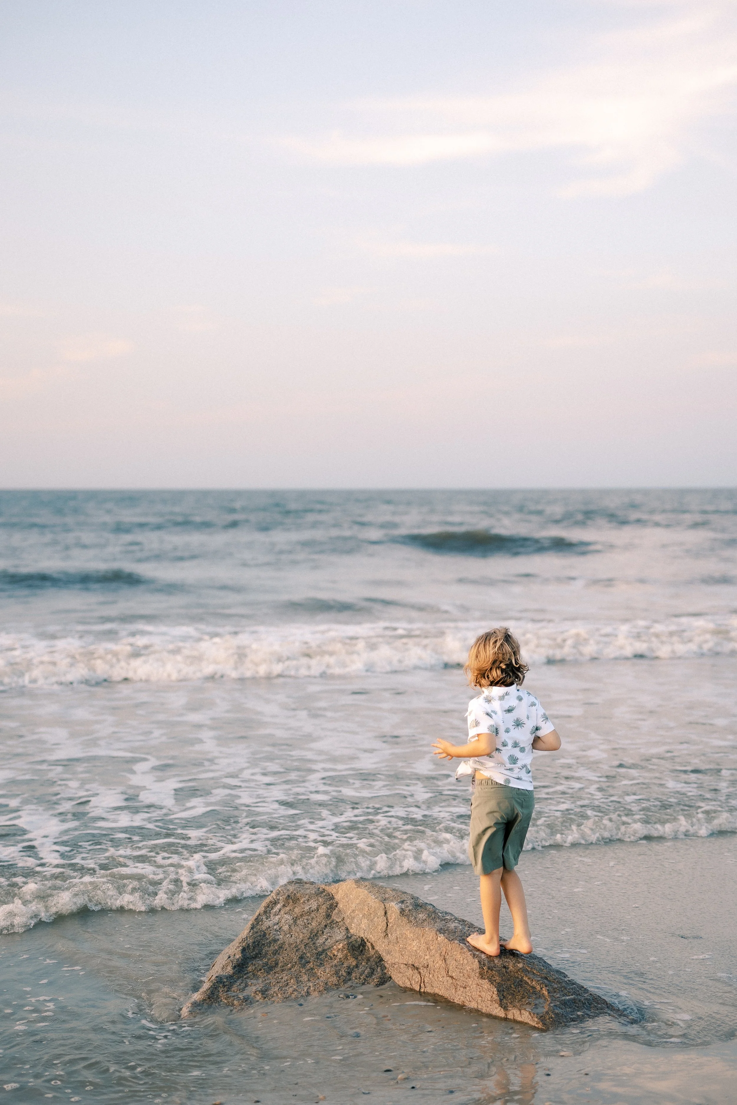 A child standing on a large rock at the beach, looking at the ocean waves under a cloudy sky.