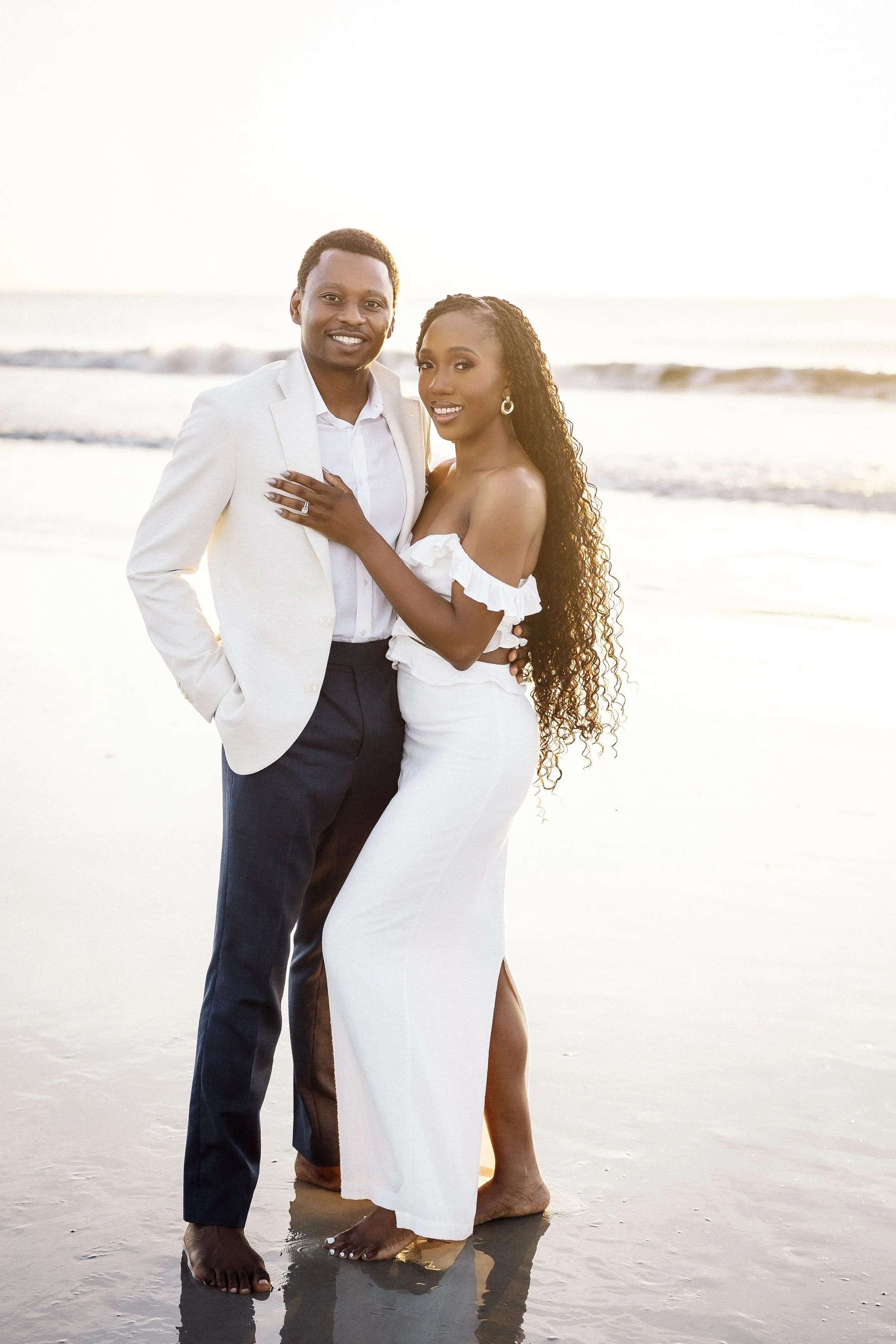 A smiling couple standing on the beach at sunset, with the ocean in the background. The man is wearing a white blazer and dark pants, and the woman is dressed in a white off-the-shoulder dress. They are barefoot, standing on wet sand, and appear happ