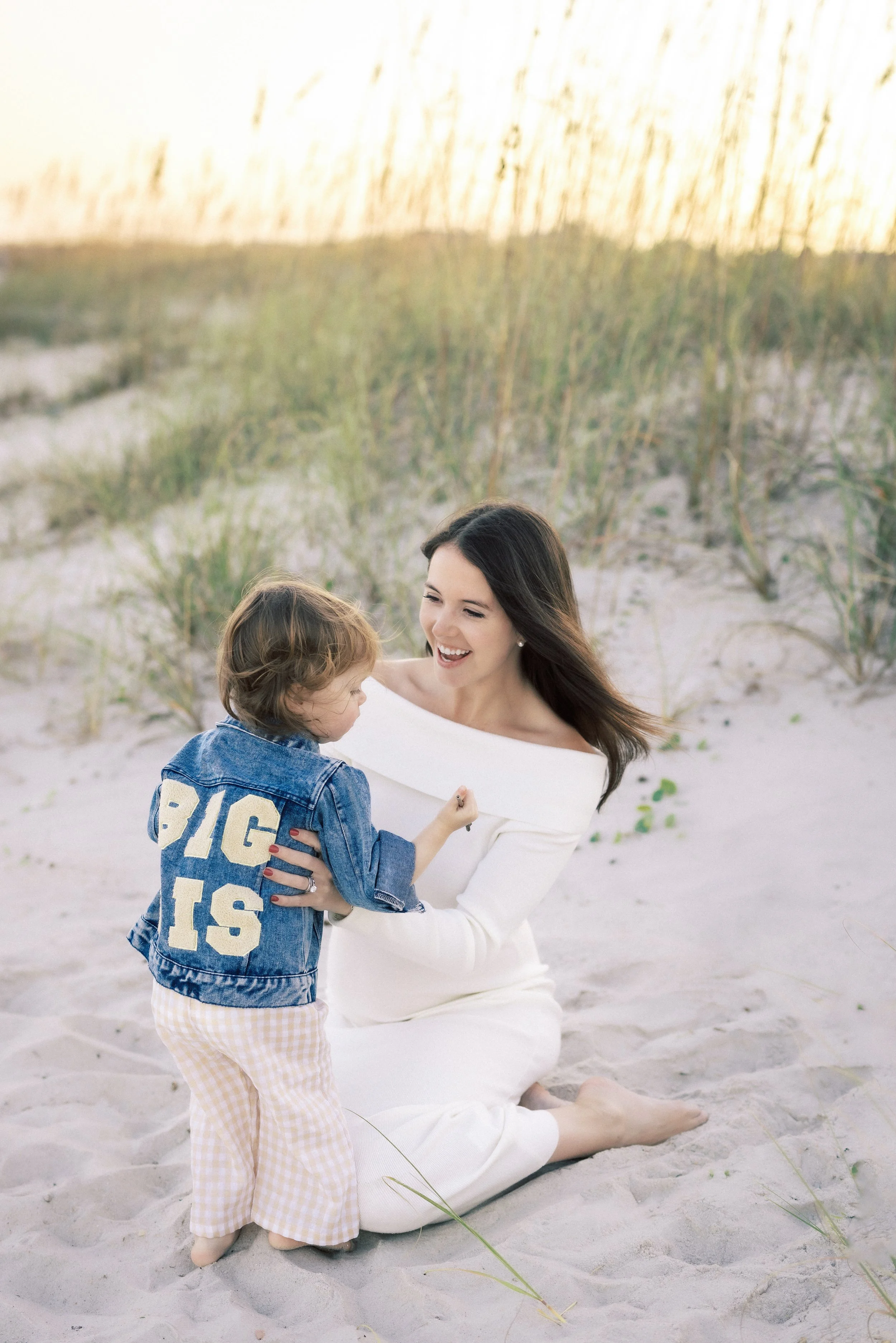 A woman in a white dress kneeling on the sand with a young boy in a denim jacket and yellow checkered pants.Photographer Amelia Island, Jacksonville, St Augustine, St Simons Island, Savannah photographer