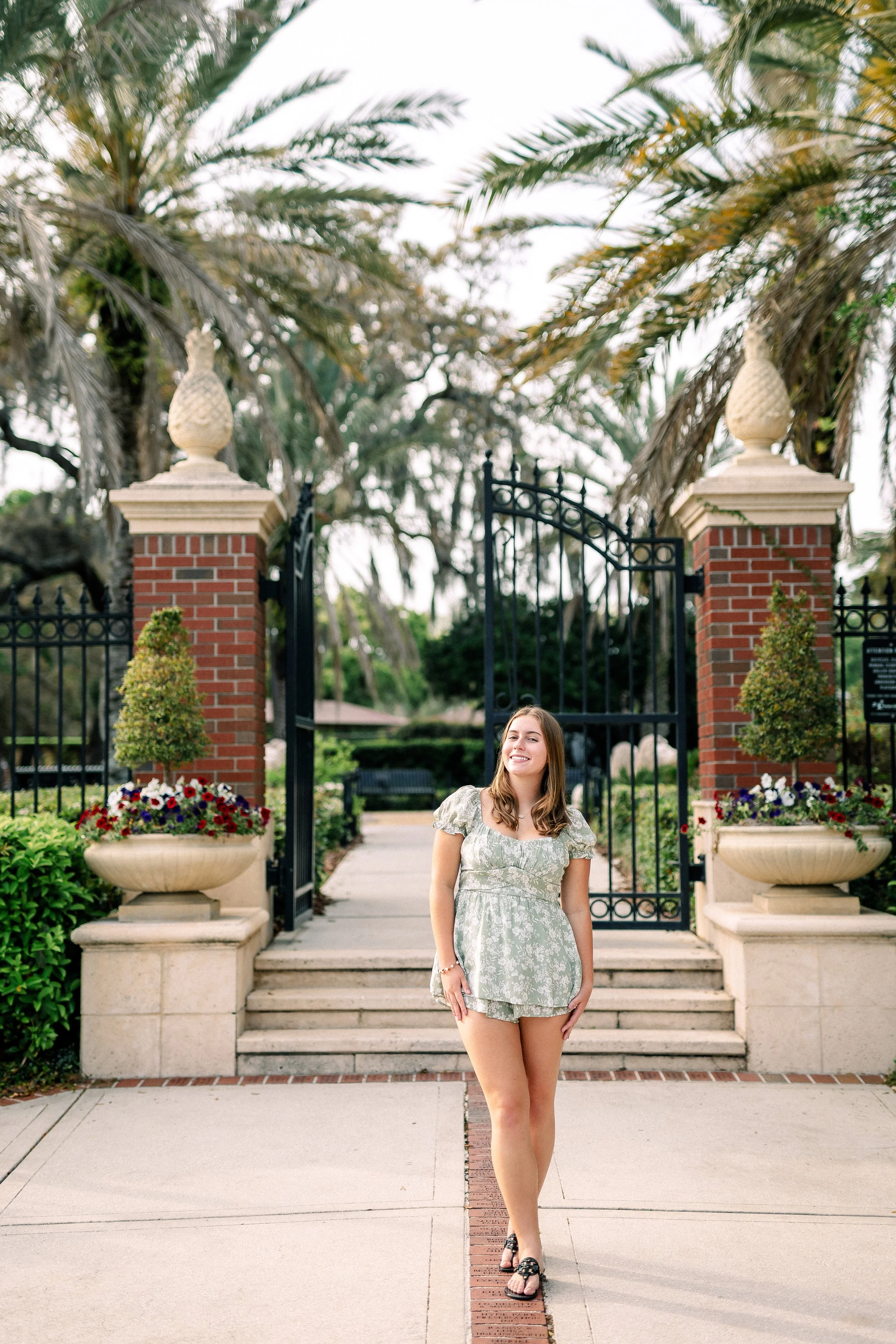 A young woman with long brown hair, wearing a light green floral dress and black sandals, is standing on a sidewalk in front of a decorative iron gate with brick pillars, all surrounded by lush greenery and large potted flowers.