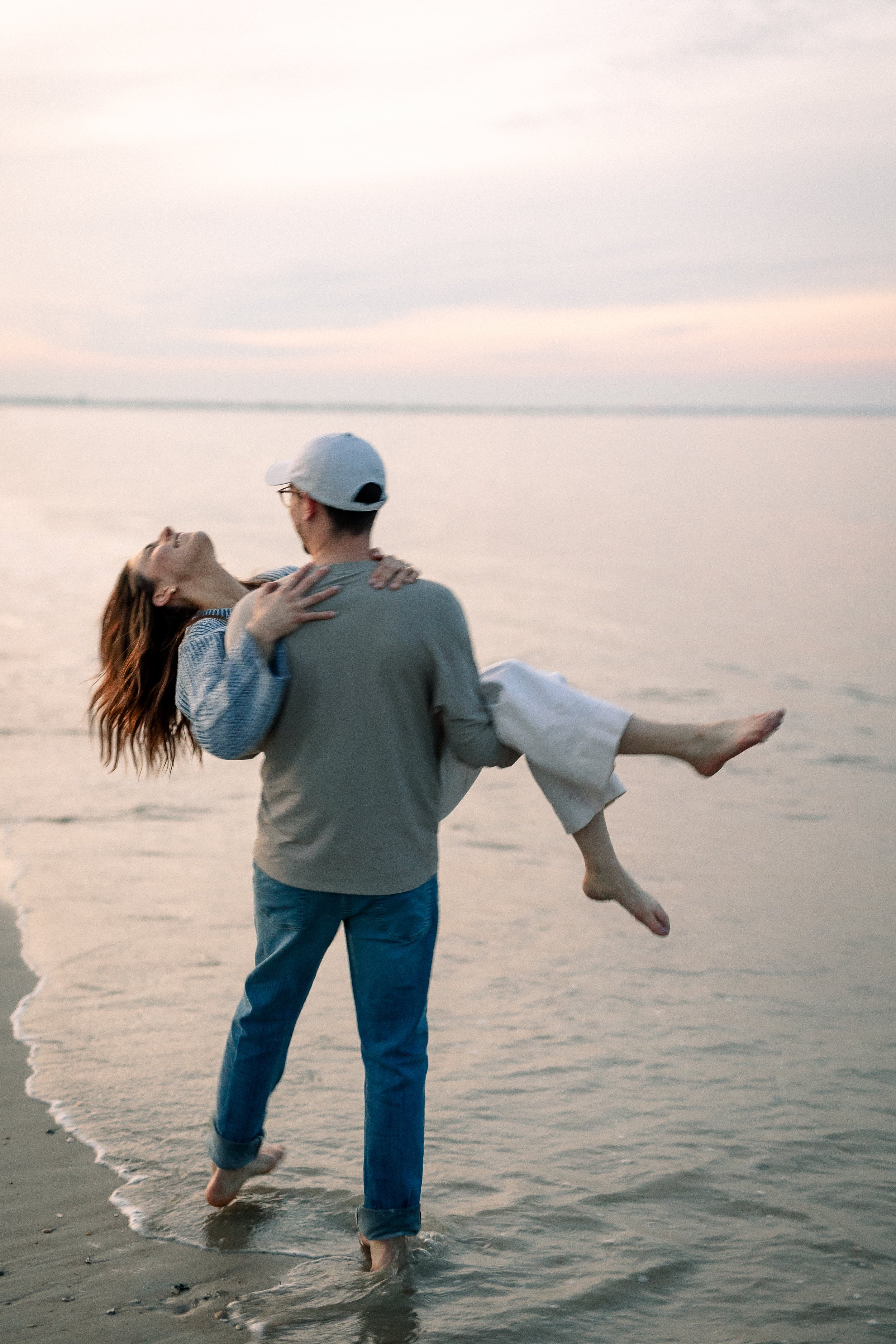 A man is holding a woman in his arms at the beach during sunset, with the water and sky visible in the background.