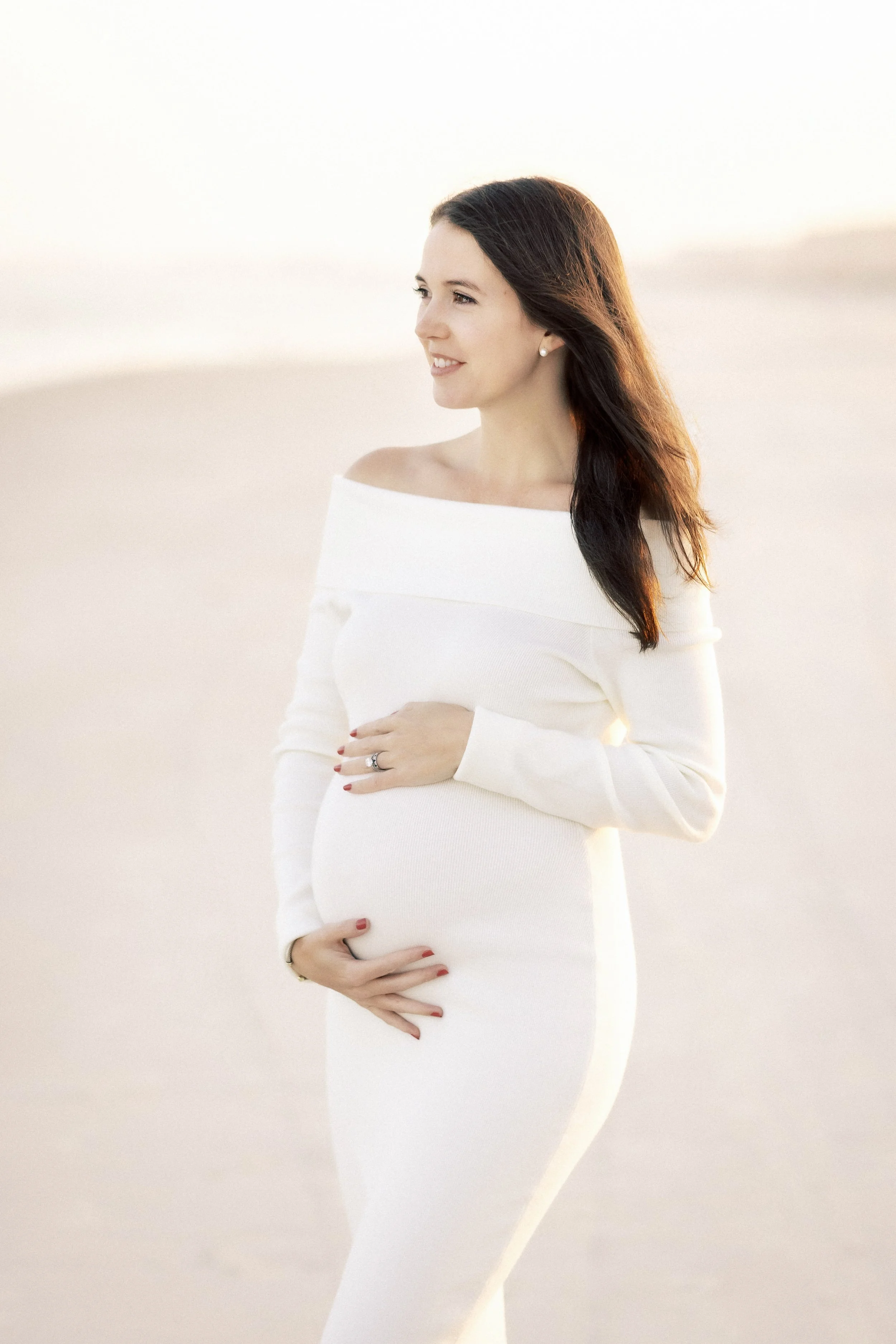 A pregnant woman in a white dress standing outdoors on a beach at sunset, cradling her belly with her hands.Photographer Amelia Island, Jacksonville, St Augustine, St Simons Island, Savannah photographer