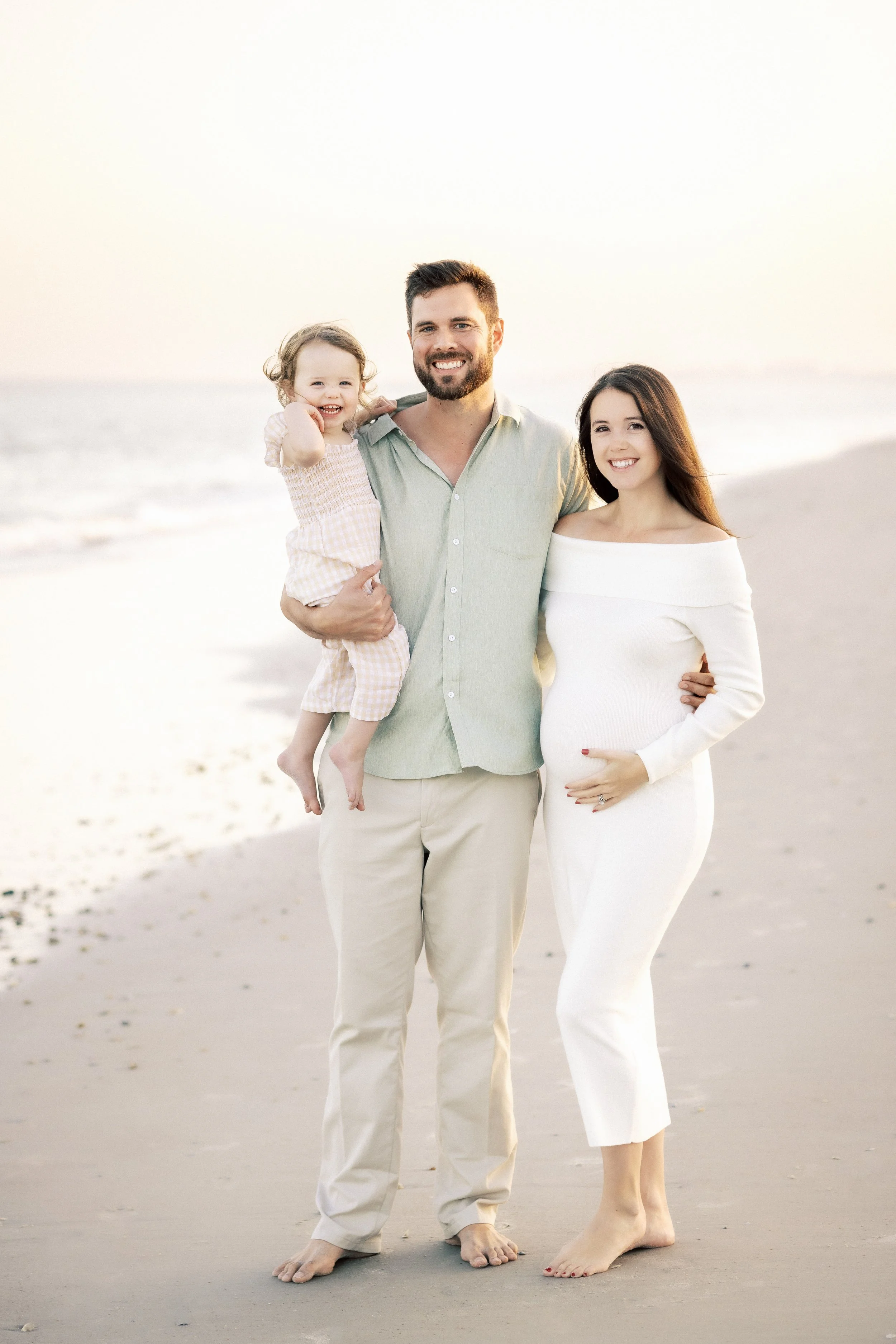 A family of three standing on a beach during sunset. The father, holding their young daughter, is smiling and wearing a light-colored shirt and beige pants. Photographer Amelia Island, Jacksonville, St Augustine, St Simons Island, Savannah photograph