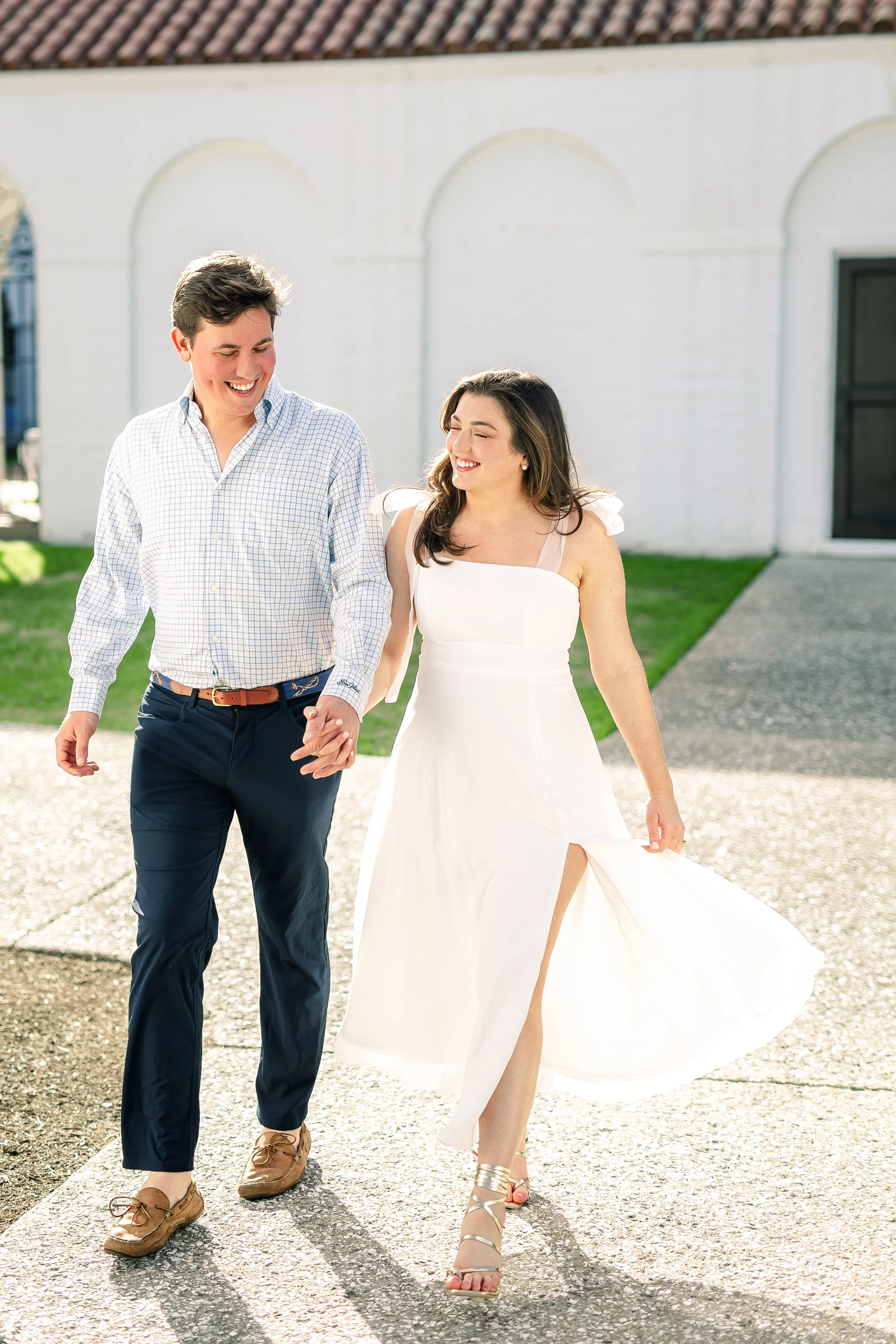 A young couple walking outdoors hand in hand, smiling and looking at each other. The woman is wearing a white dress with slit and high-heeled sandals, while the man is dressed in a checkered shirt and dark trousers with brown shoes. The background fe