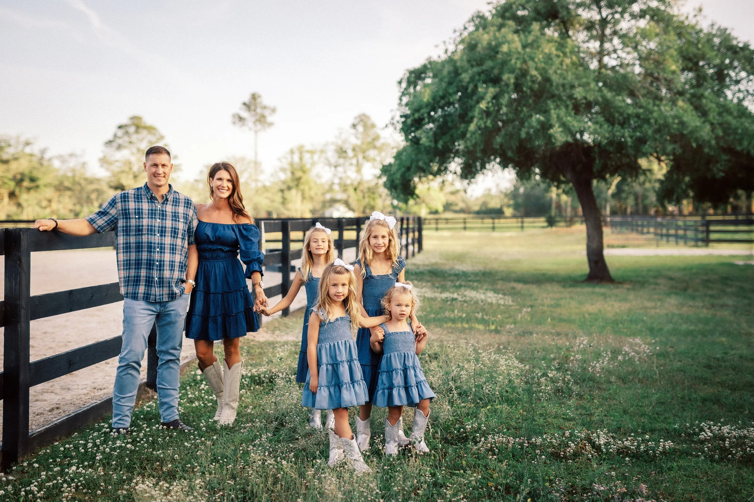 A family of six standing outdoors near a black fence in a grassy area with a large tree, all smiling and holding hands. The family includes two adults and four young girls, some of whom are wearing matching blue dresses and cowboy boots.