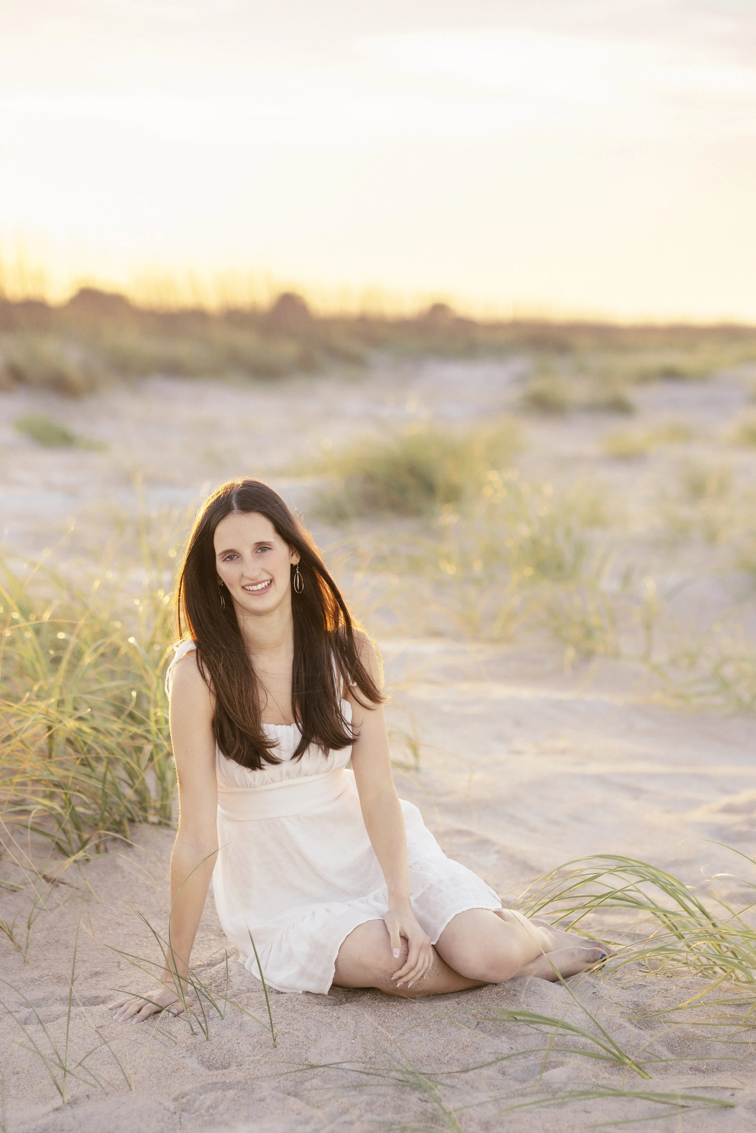 A woman with long dark hair sitting on a sandy beach surrounded by grass, wearing a white dress, smiling at the camera during sunset.