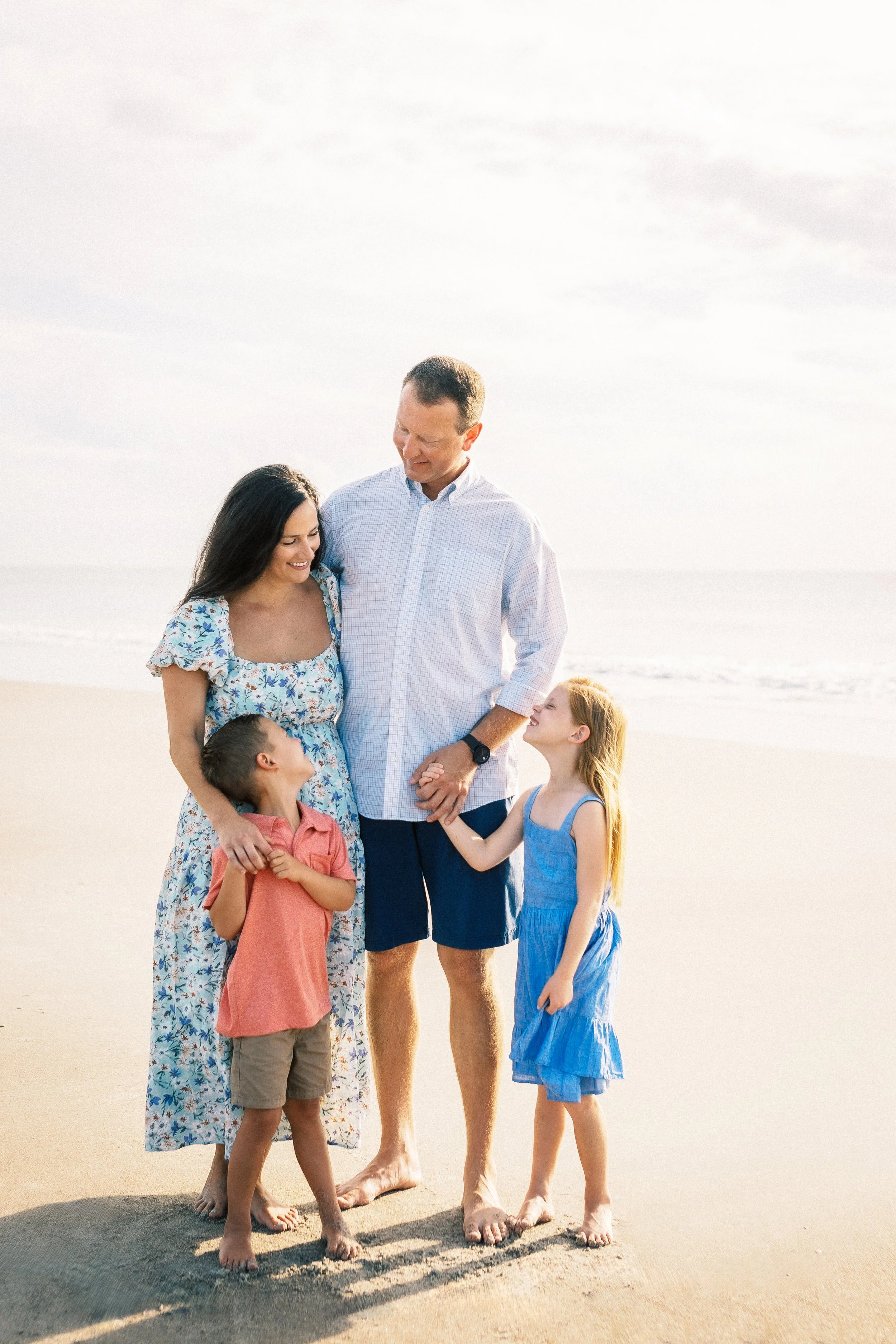 A family of six standing barefoot on a sandy beach, smiling and looking at each other, with the ocean and sky in the background.Photographer Amelia Island, Jacksonville, St Augustine, St Simons Island, Savannah photographer