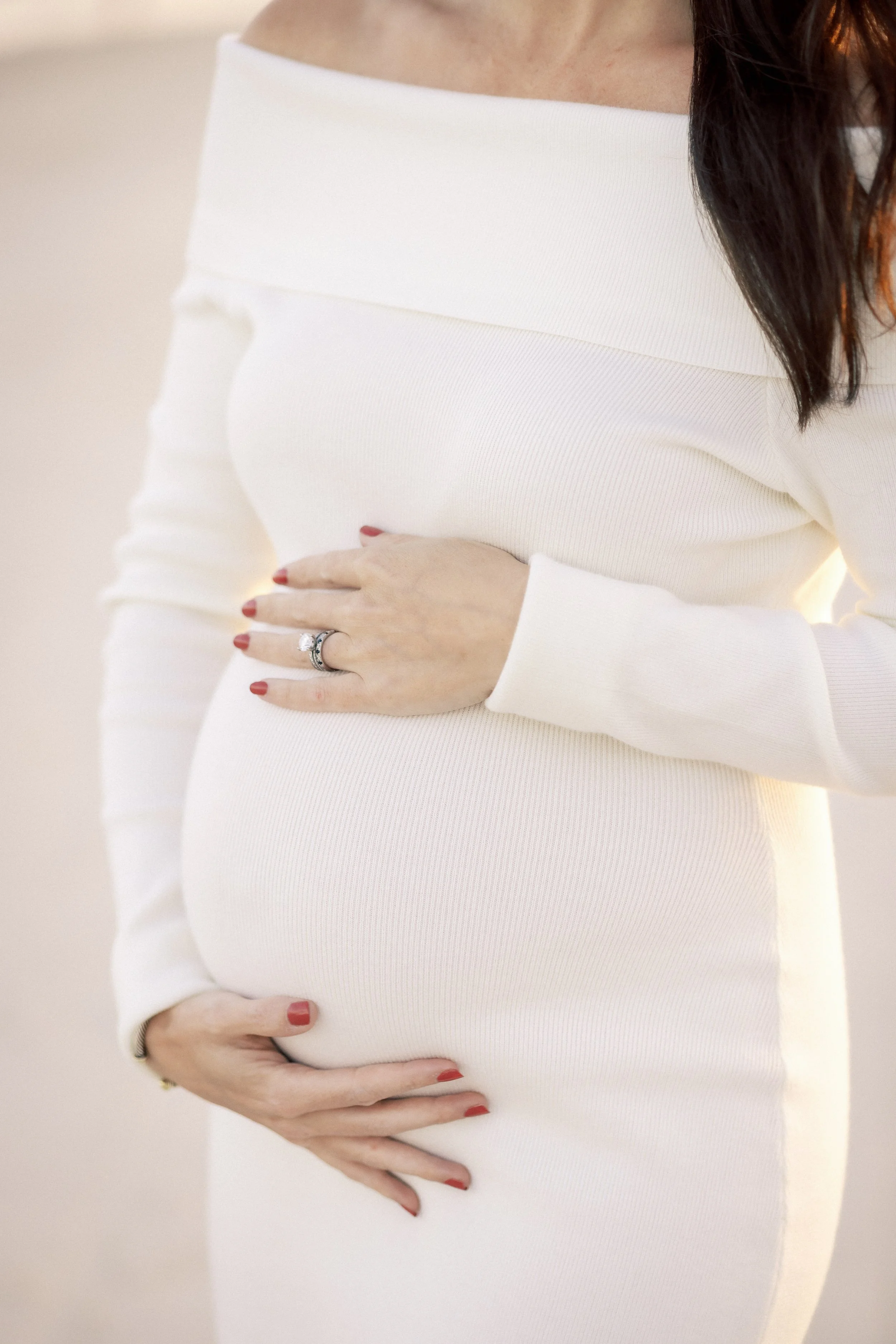 A woman in a white long-sleeve dress is holding her pregnant belly with both hands. She is wearing a ring on her left hand and has red-painted nails.Photographer Amelia Island, Jacksonville, St Augustine, St Simons Island, Savannah photographer