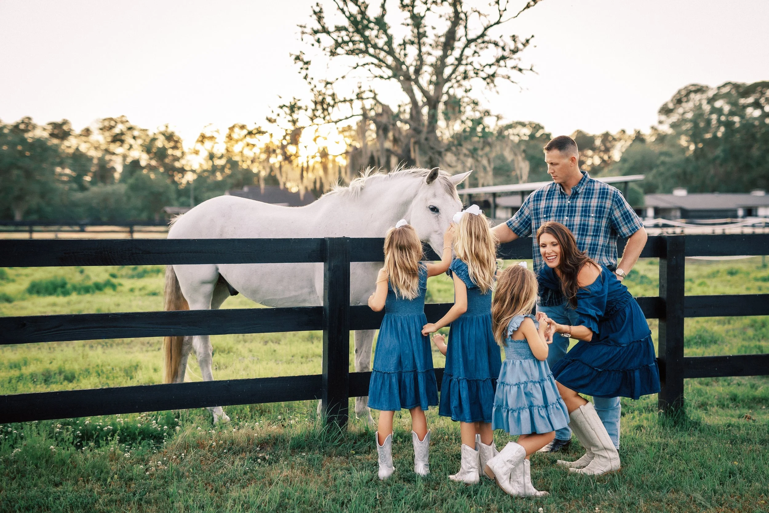 A group of four young girls in blue dresses and white boots, a woman, and a man interacting with a white horse behind a black fence in a green field during sunset, with trees in the background.