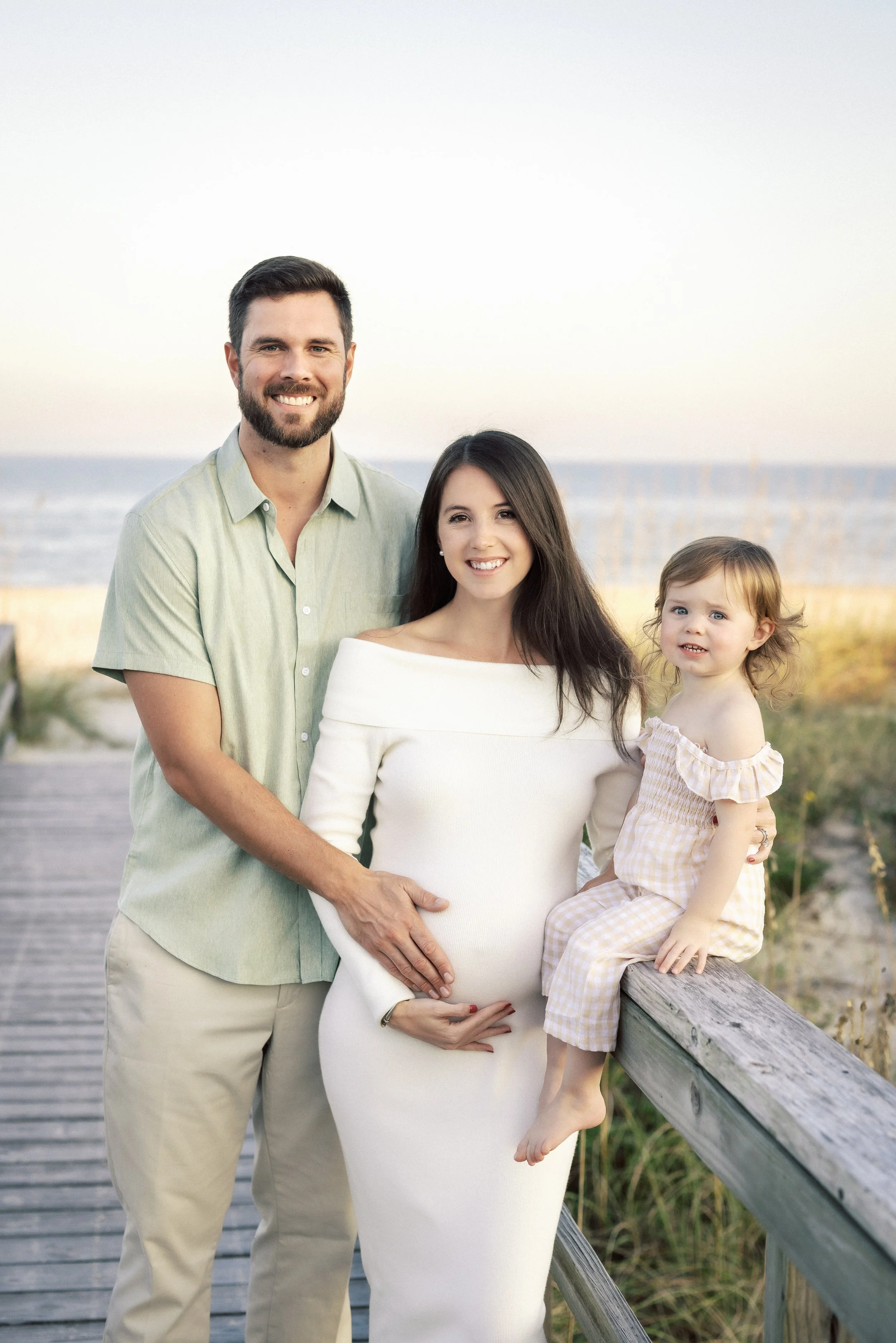 A smiling family of three standing on a wooden boardwalk near the beach, with a man, woman, and young girl, during sunset.Photographer Amelia Island, Jacksonville, St Augustine, St Simons Island, Savannah photographer