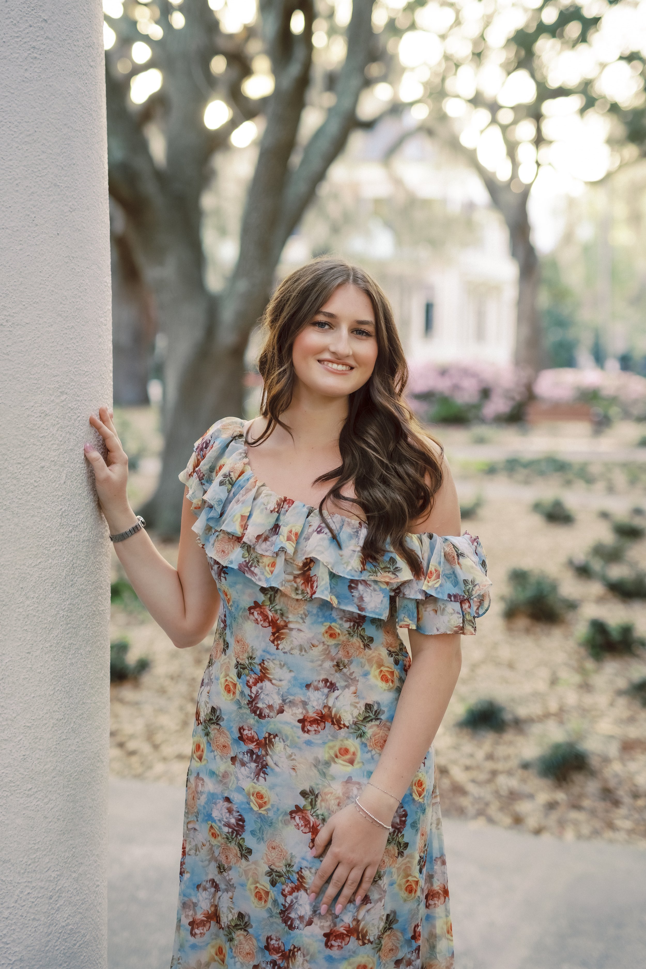 A young woman with long wavy brown hair wearing a floral dress, standing outdoors next to a white column, smiling, with trees and a park in the background during daytime.