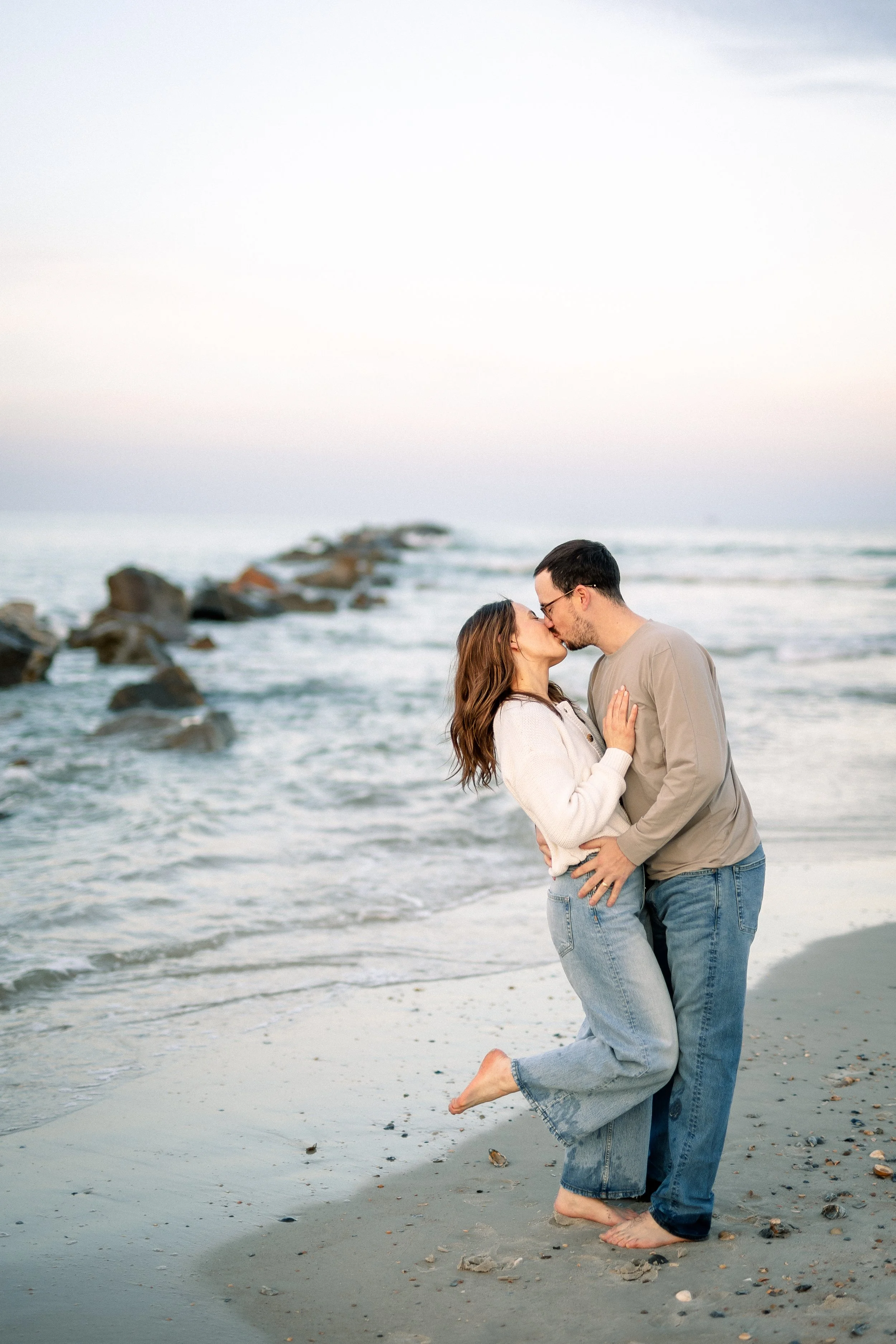 A couple kissing on the beach, with the man kneeling on one knee and the woman holding his face, both with their eyes closed. The ocean waves and rocks are in the background.