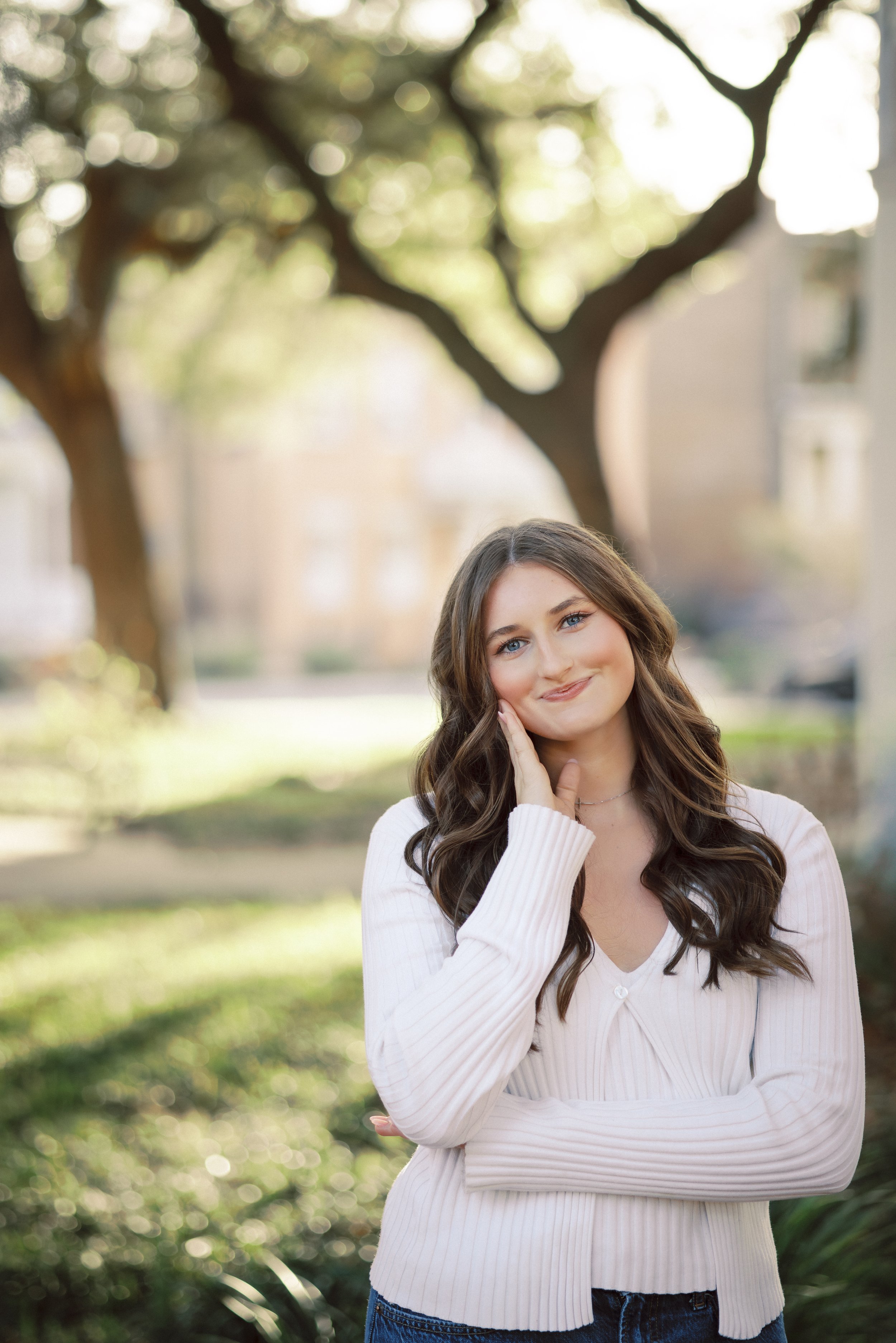Close-up of a young woman with long wavy brown hair, blue eyes, and fair skin, standing outdoors in front of a large tree and green shrubs, smiling softly and touching her face with one hand, wearing a white ribbed sweater and blue jeans.