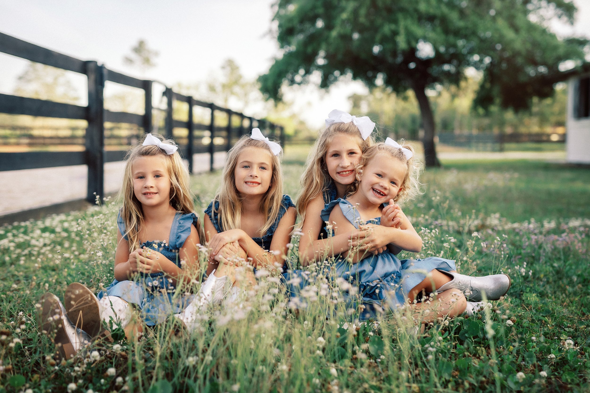 Four young girls sitting together in a grassy field full of small white flowers, smiling at the camera. They are wearing blue dresses with white bows in their hair and appear to be enjoying a day outdoors on a sunny day. In the background, there are 