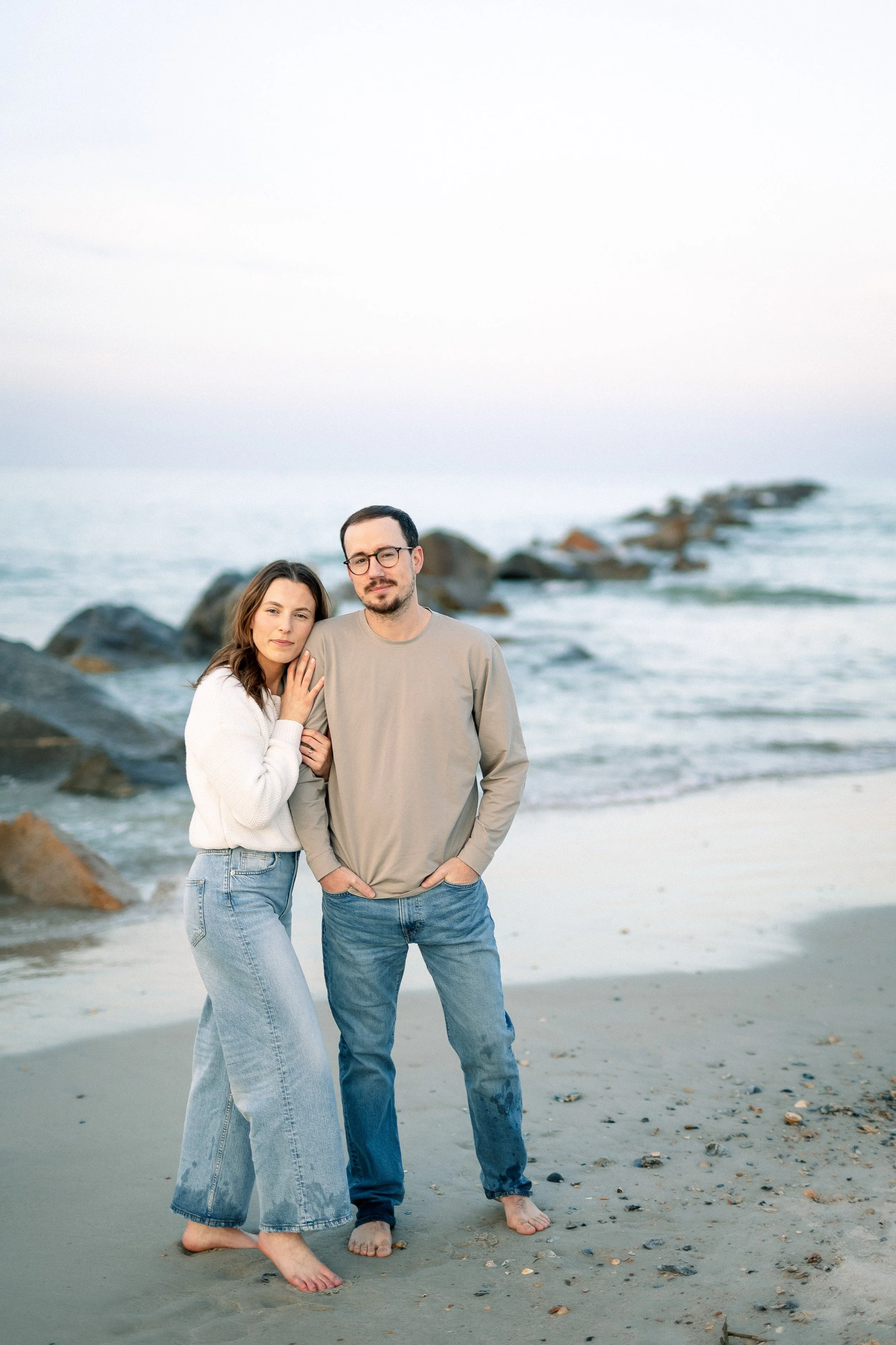 A young woman and a young man standing on the beach with rocks and ocean in the background. They are barefoot and close together, with the woman touching the man's shoulder. The woman has long brown hair, and the man has short dark hair, glasses, and