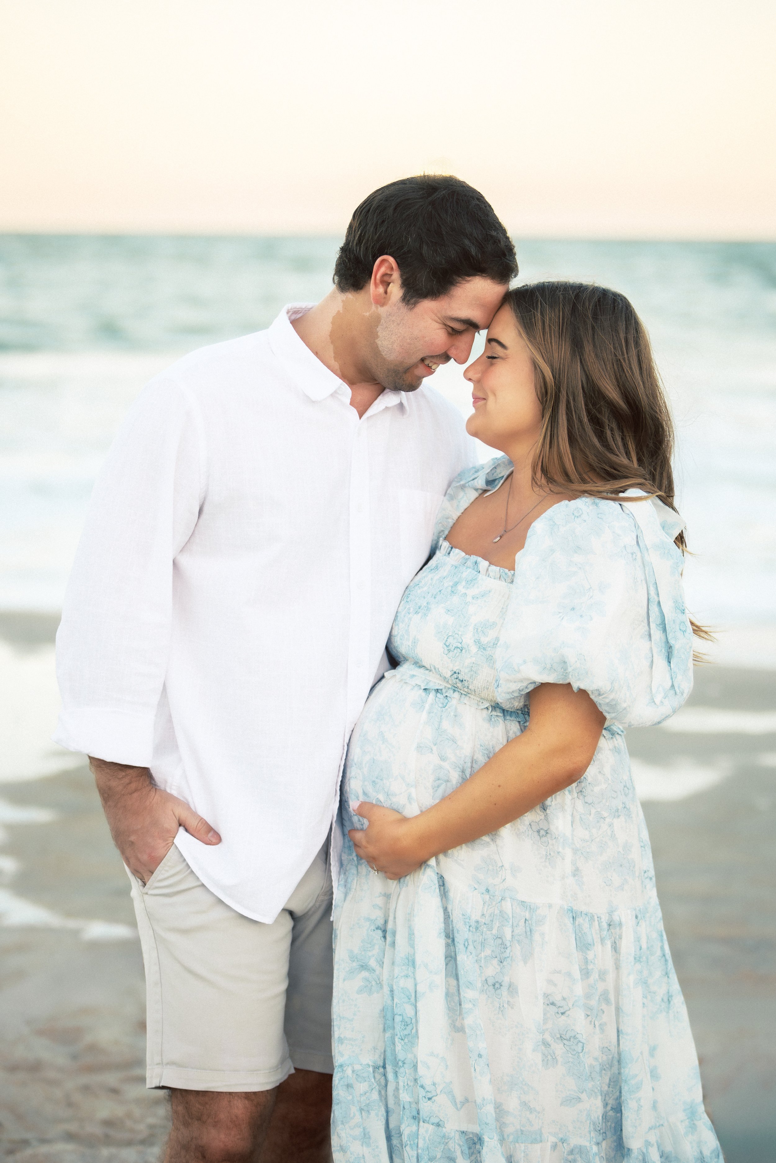 A couple standing close on the beach with foreheads touching, eyes closed, smiling, with ocean waves in the background.