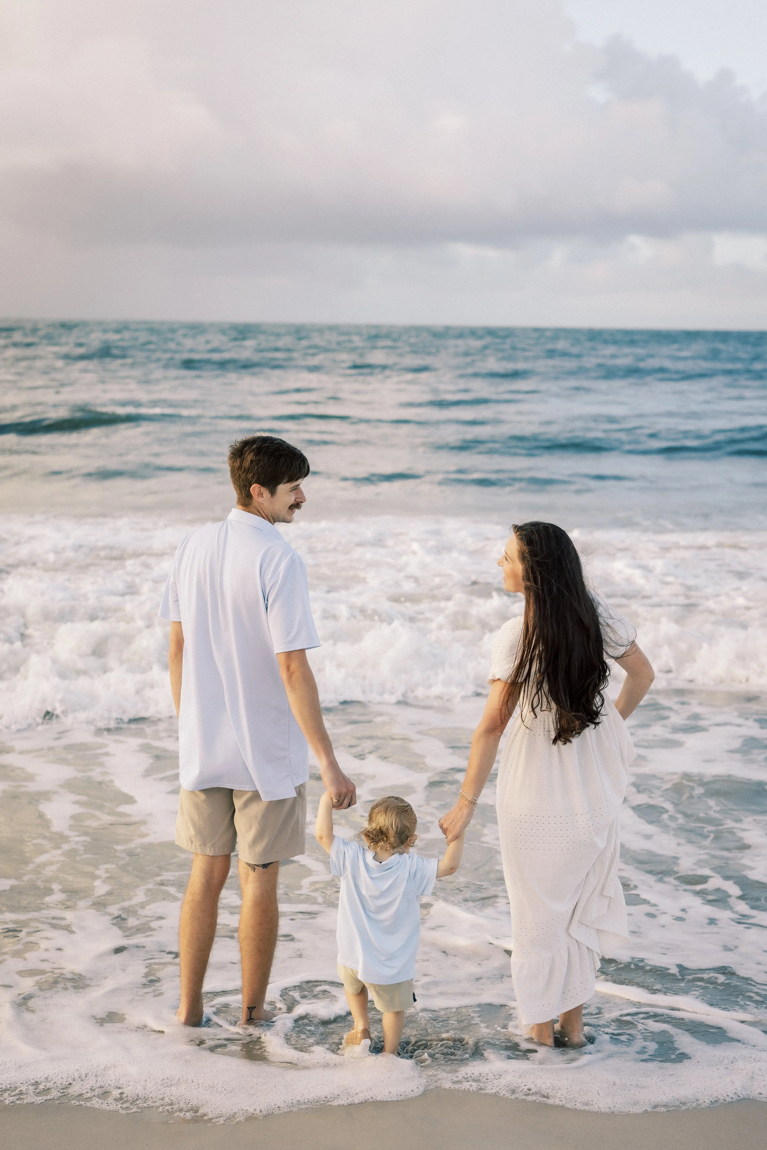 A family of four at the beach, including a man, woman, and two young children, holding hands and standing in the shallow surf with cloudy skies overhead.Photographer Amelia Island, Jacksonville, St Augustine, St Simons Island, Savannah photographer