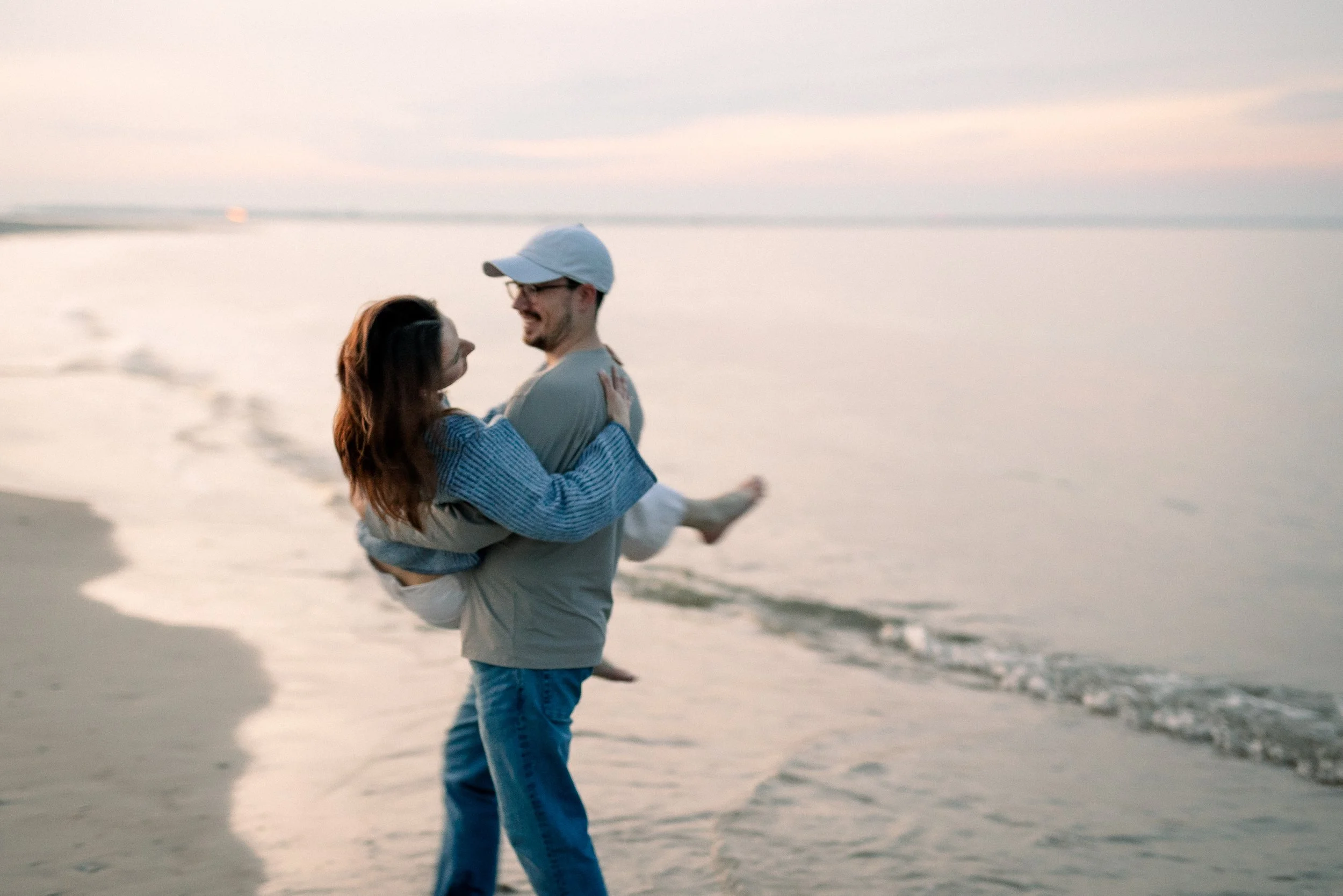 A couple dancing on the beach during sunset, with the man lifting the woman in his arms, the ocean in the background.