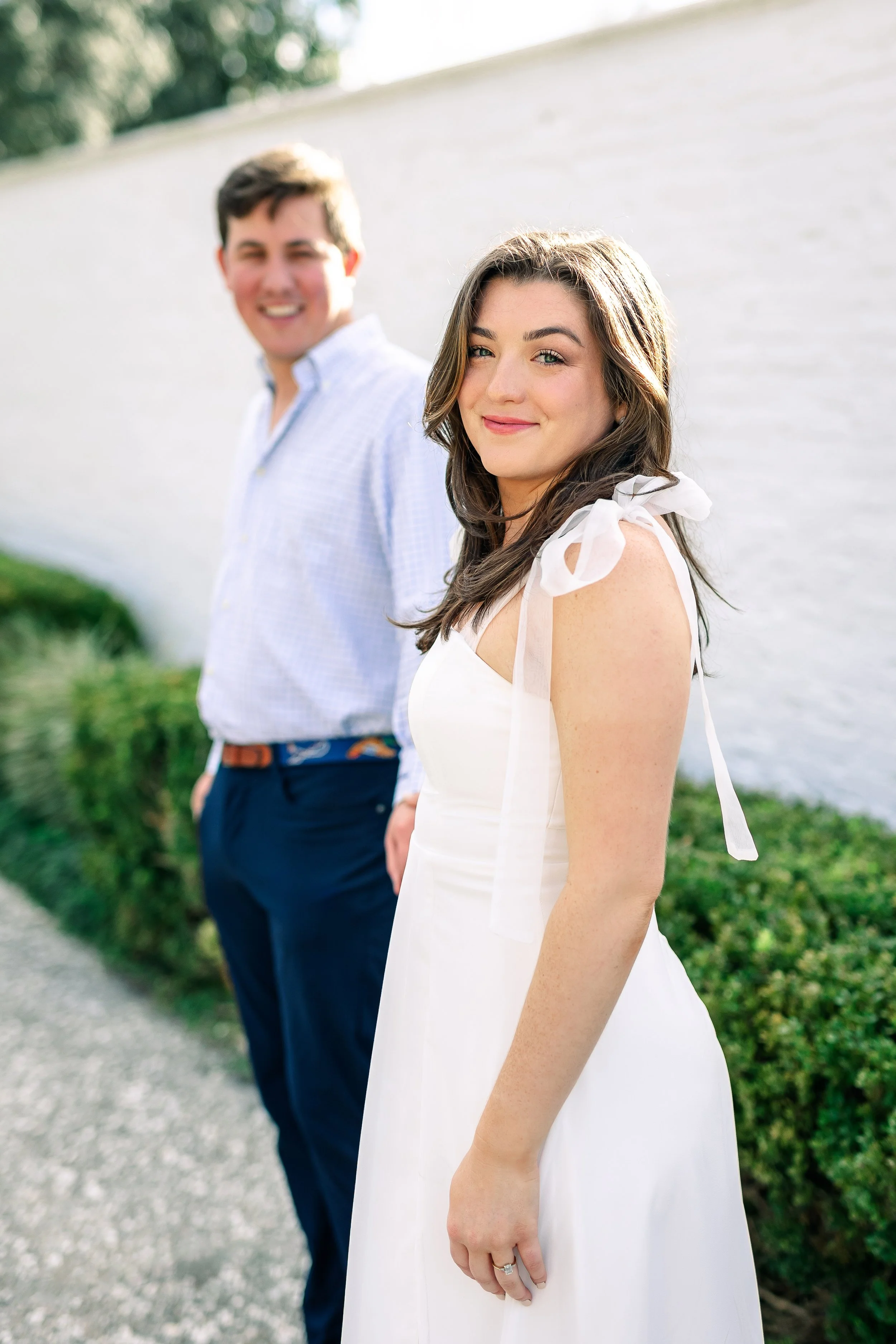 A young woman with brown hair and blue eyes smiling at the camera, wearing a white dress with shoulder ribbons, standing outdoors near a white wall, with a young man with dark hair and a white checkered shirt blurred in the background.