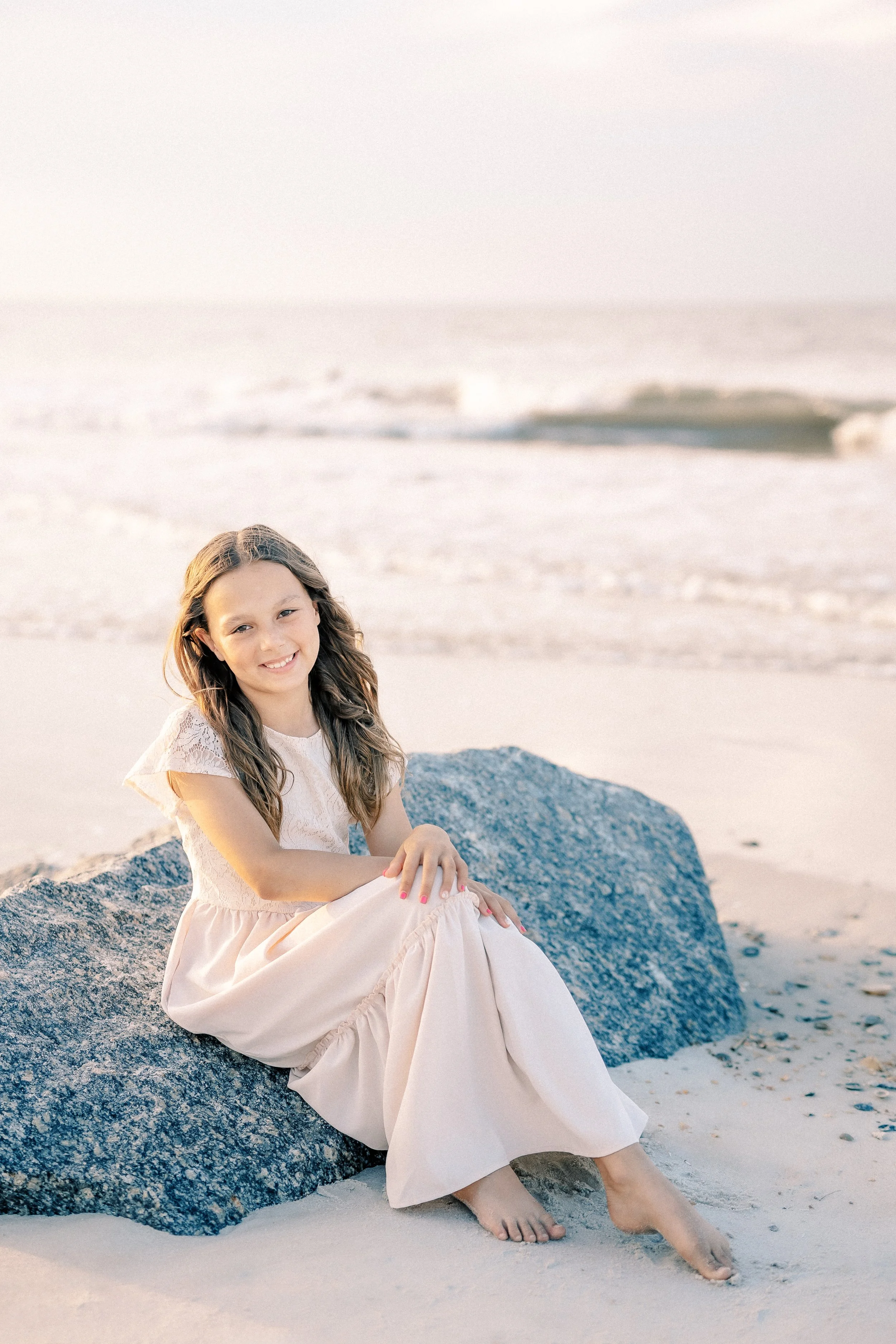 A young girl with long, wavy brown hair sitting on a large rock at the beach, wearing a light pink dress with lace details, smiling, with waves and sandy shore in the background.