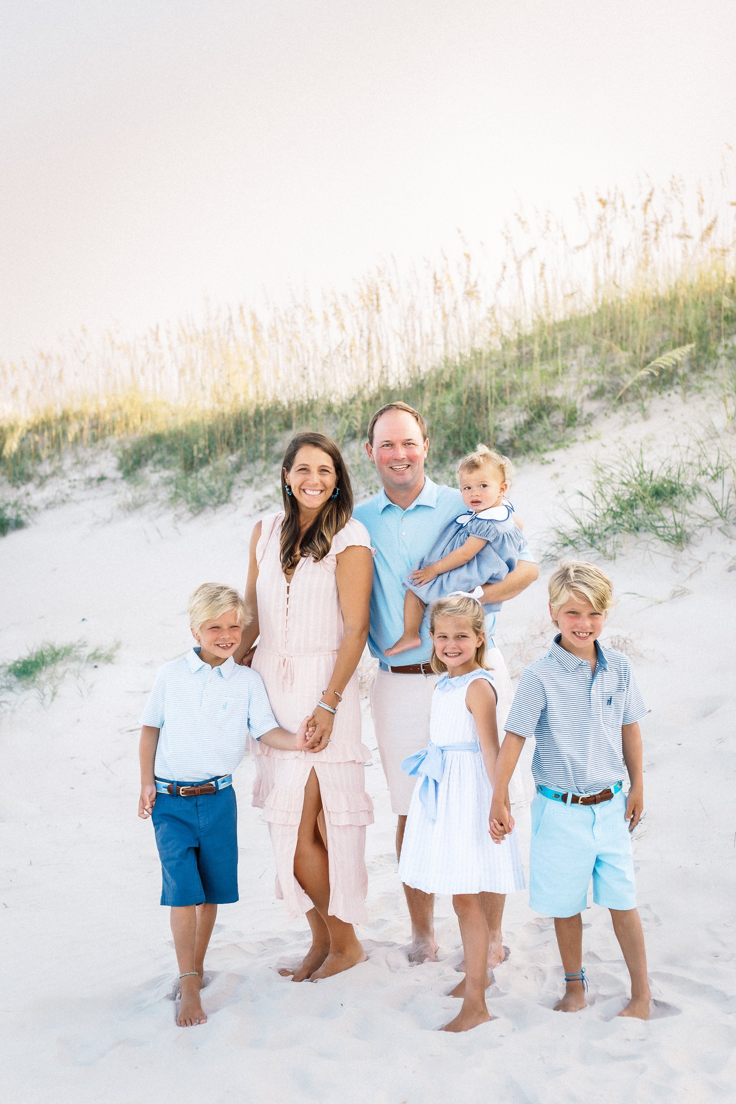 A family walking on a sandy beach with grass-covered dunes in the background. The family members are wearing light, summery clothing and smiling at the camera.Photographer Amelia Island, Jacksonville, St Augustine, St Simons Island, Savannah photogra