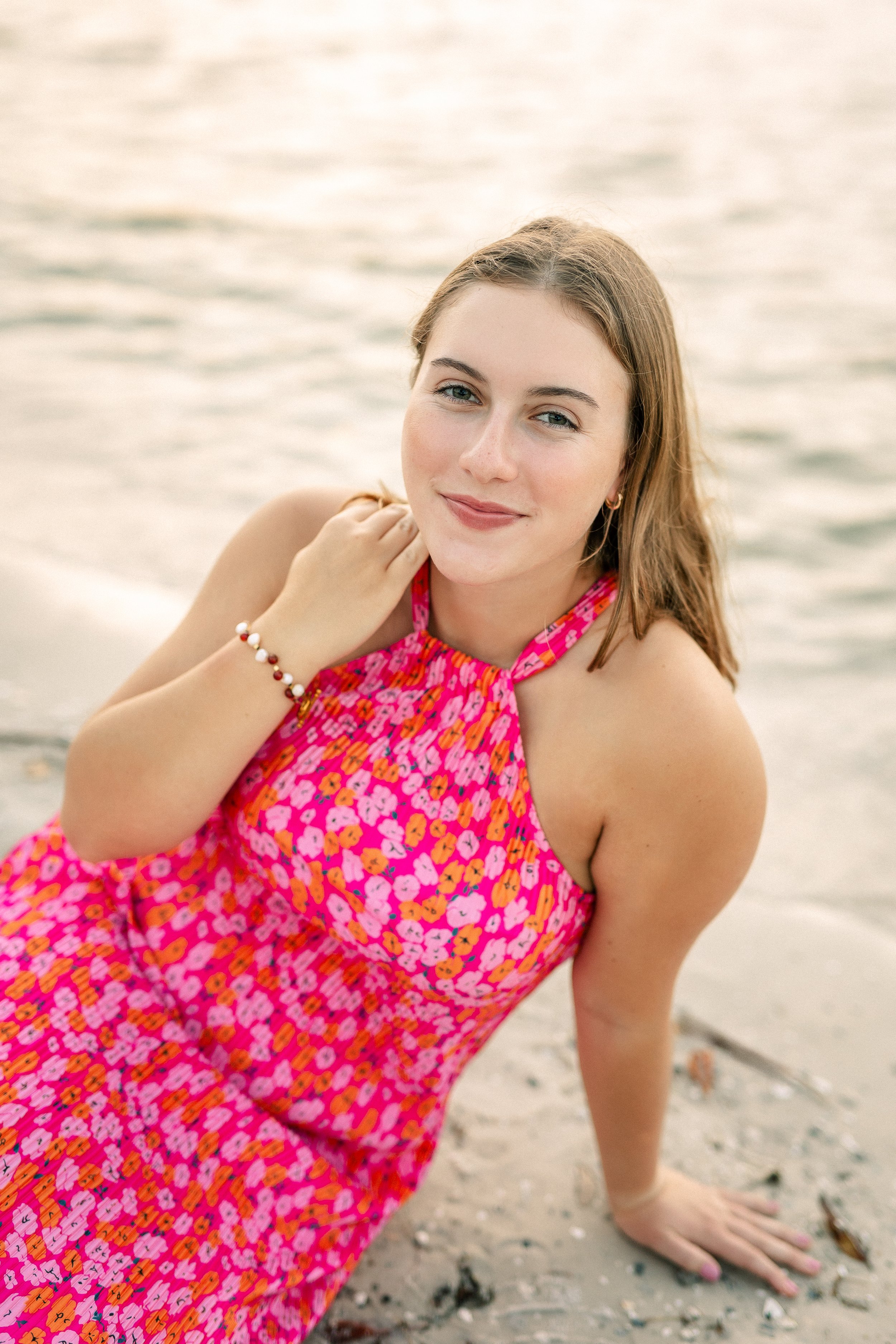 Young woman with light brown hair, blue eyes, and fair skin, wearing a pink floral dress, sitting on sandy beach near water, smiling at the camera with her hand resting on her shoulder.