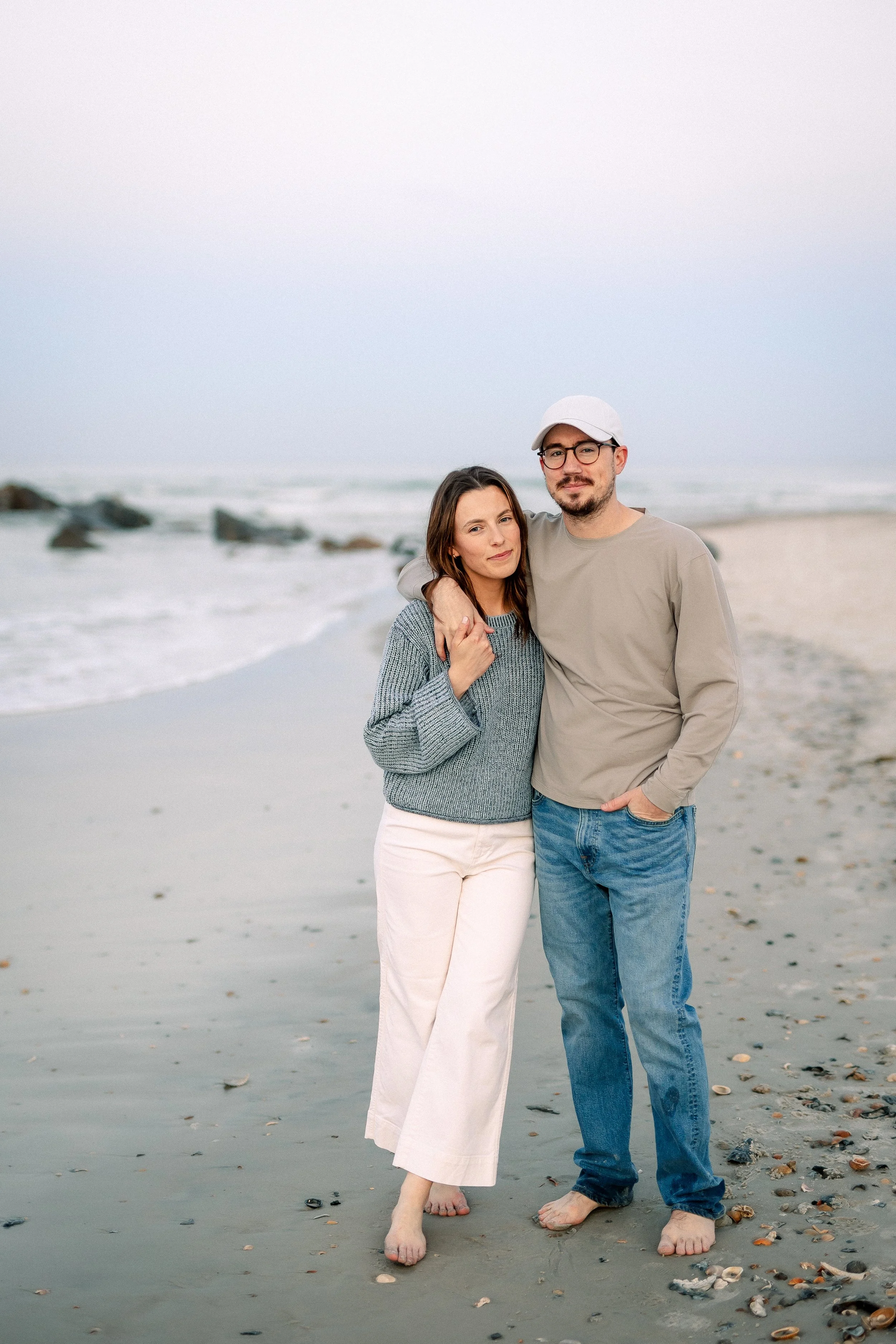 A couple standing together on a beach, the woman has brown hair and is wearing a gray sweater and white pants, and the man has glasses, a beard, and is wearing a gray shirt, jeans, and a white cap. They are barefoot, and the sky is clear with rocks a