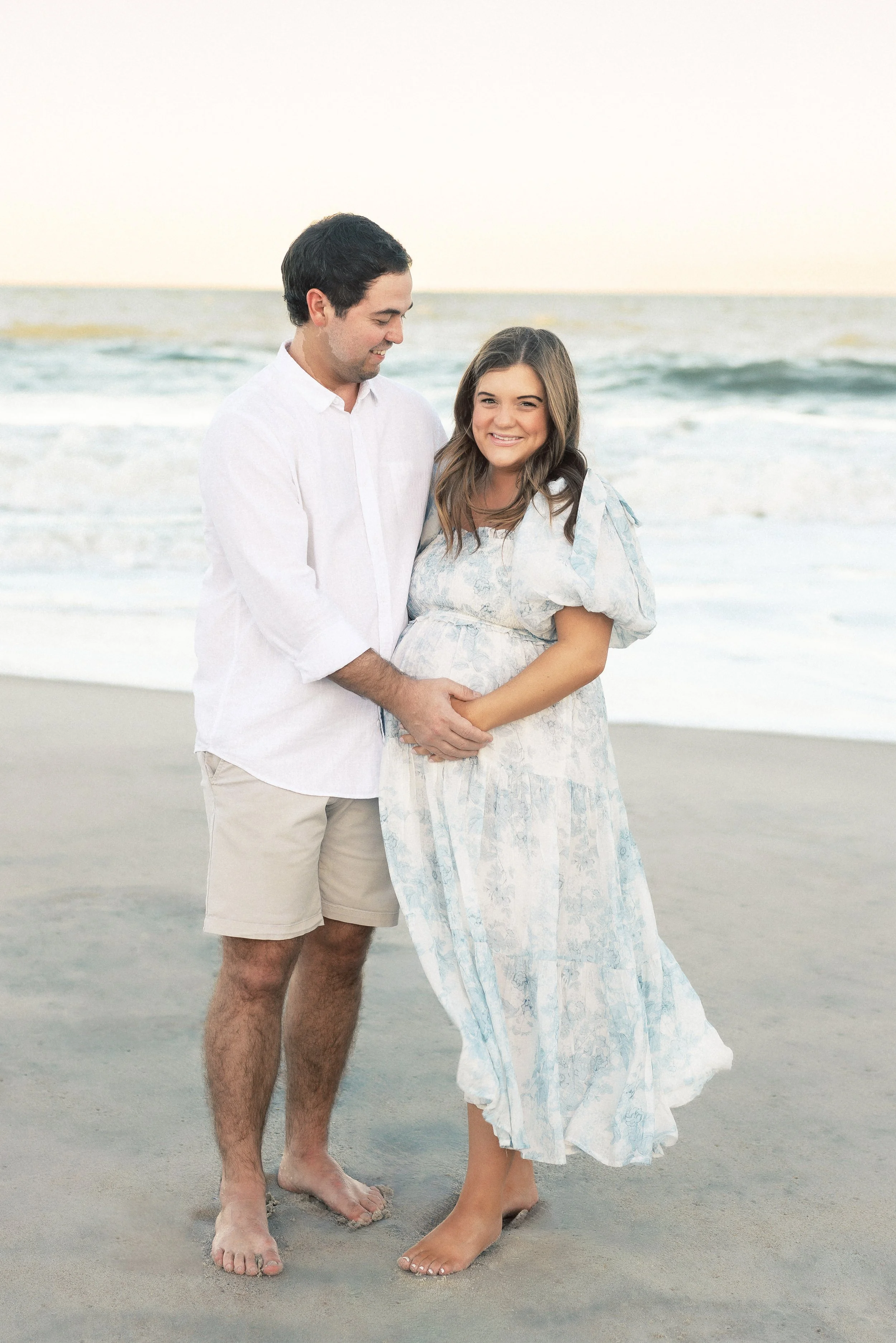 A man and a woman standing on the beach. The man is wearing a white button-up shirt and khaki shorts, smiling and looking at the woman. The woman is wearing a light blue and white dress, smiling and holding her pregnant belly. The ocean and sky are v