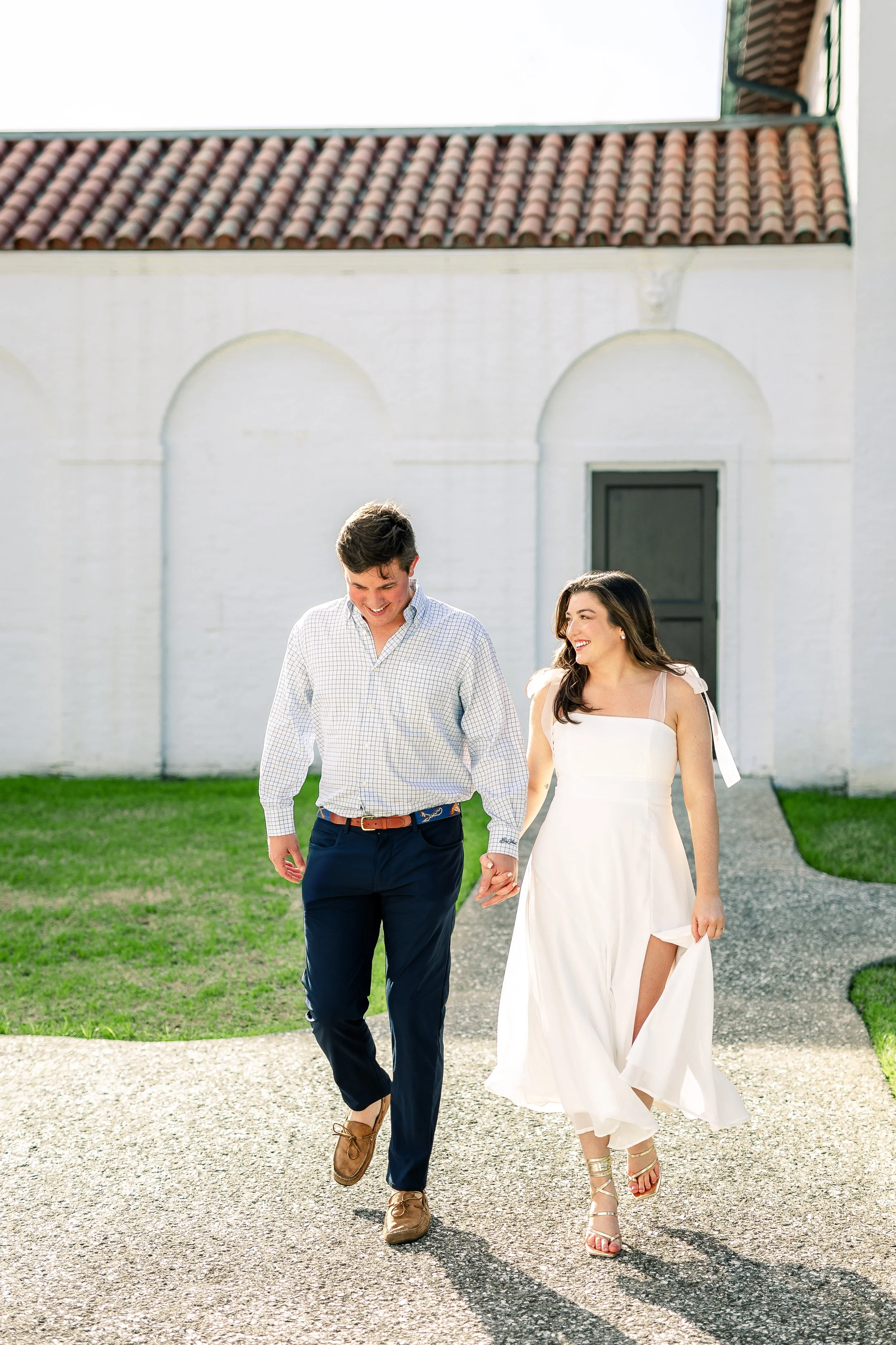 A young couple walking outdoors on a sunny day, the woman in a white dress and the man in a checkered shirt and dark pants, holding hands and smiling.