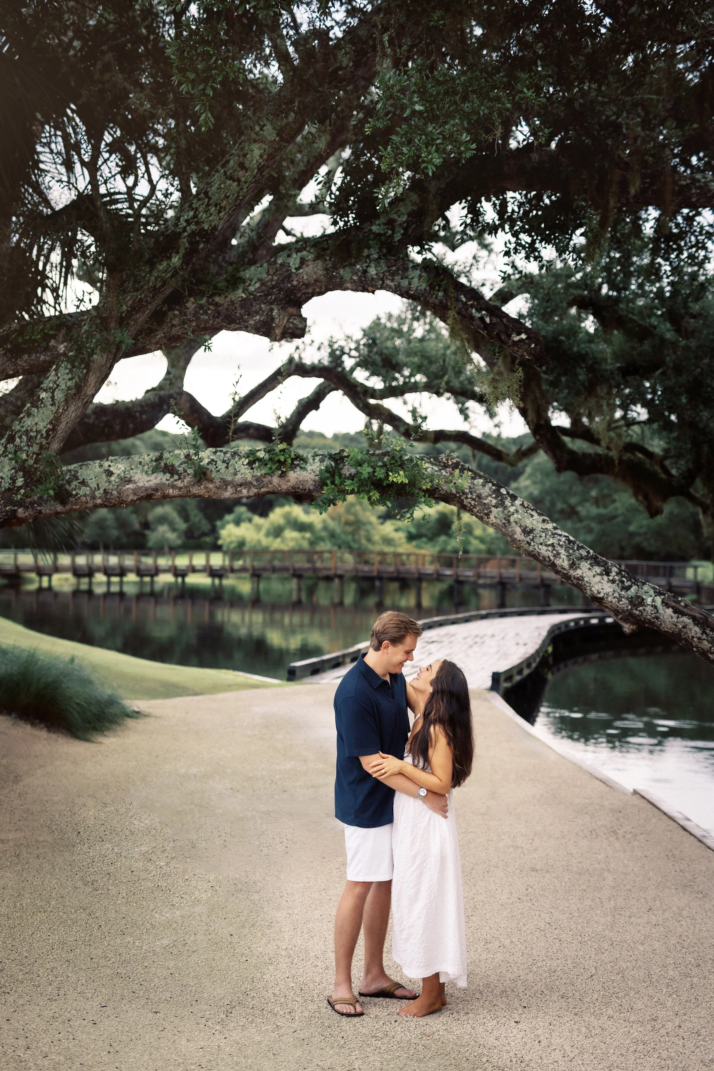A couple stands close together on a paved walkway by a calm lake, looking into each other's eyes. Behind them is a scenic landscape with a bridge over the water, lush green trees, and an overhanging large tree with twisted branches and moss. The man 