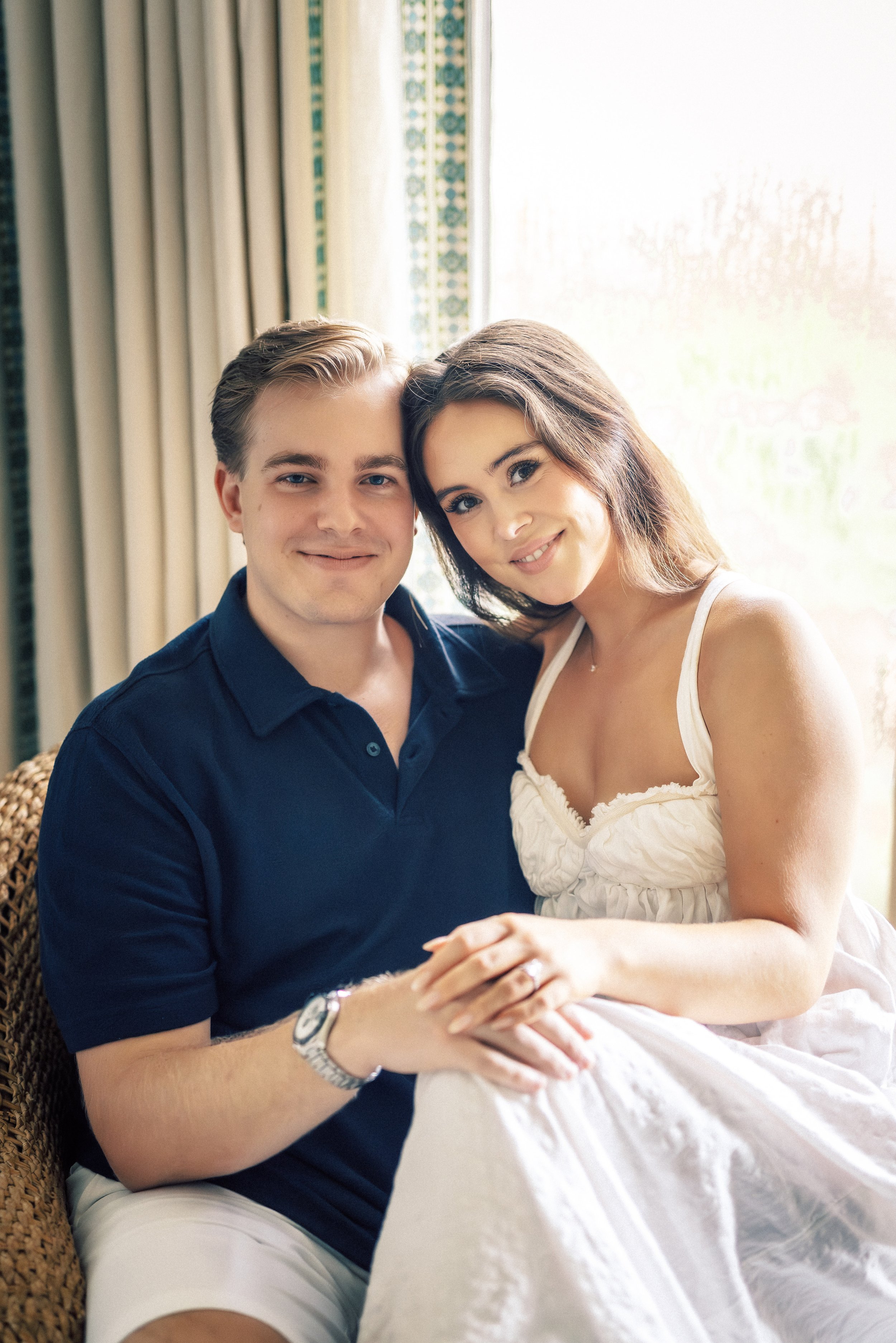 A young couple sitting close together, smiling, with the woman's head resting on the man's shoulder, in a warmly lit room with curtains and a window behind them.