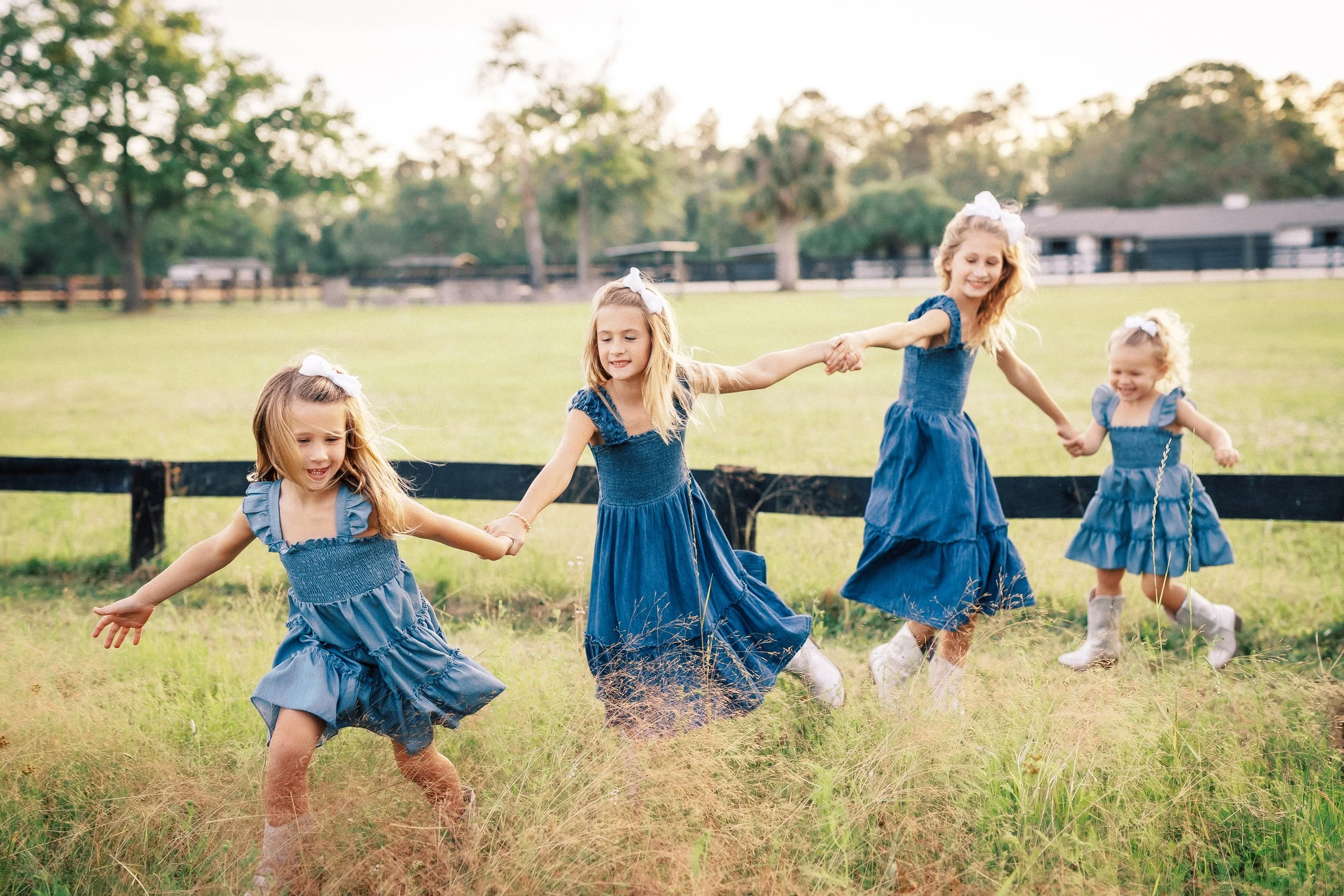 Four young girls in matching blue dresses and white bows hold hands and dance in a grassy field with trees and a fence in the background during daytime.