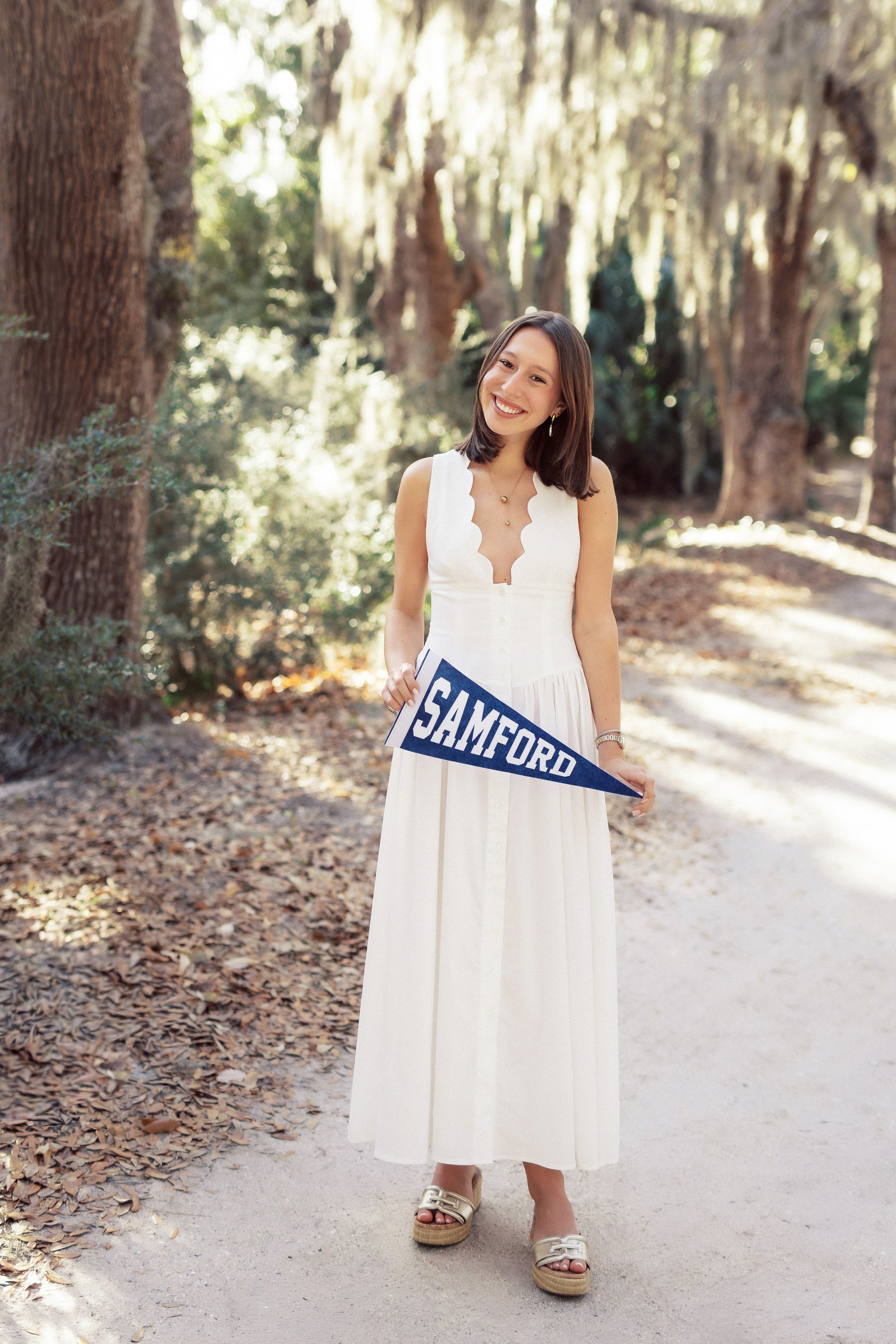 A young woman in a white dress holding a navy blue pennant that says "SAMFORD" on a wooded path during daytime.