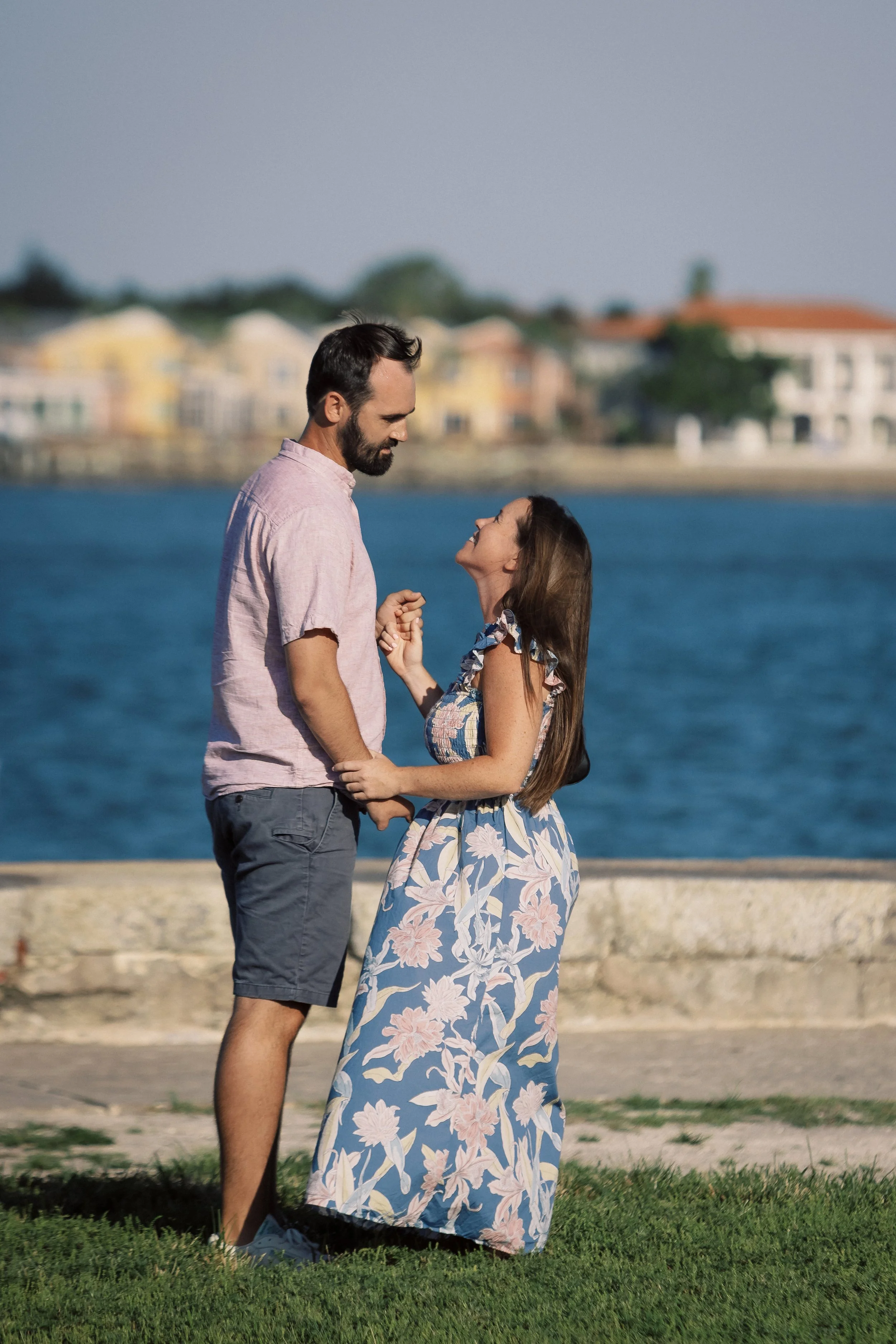 A man and a woman holding hands, looking at each other, standing outdoors near a body of water with houses in the background.