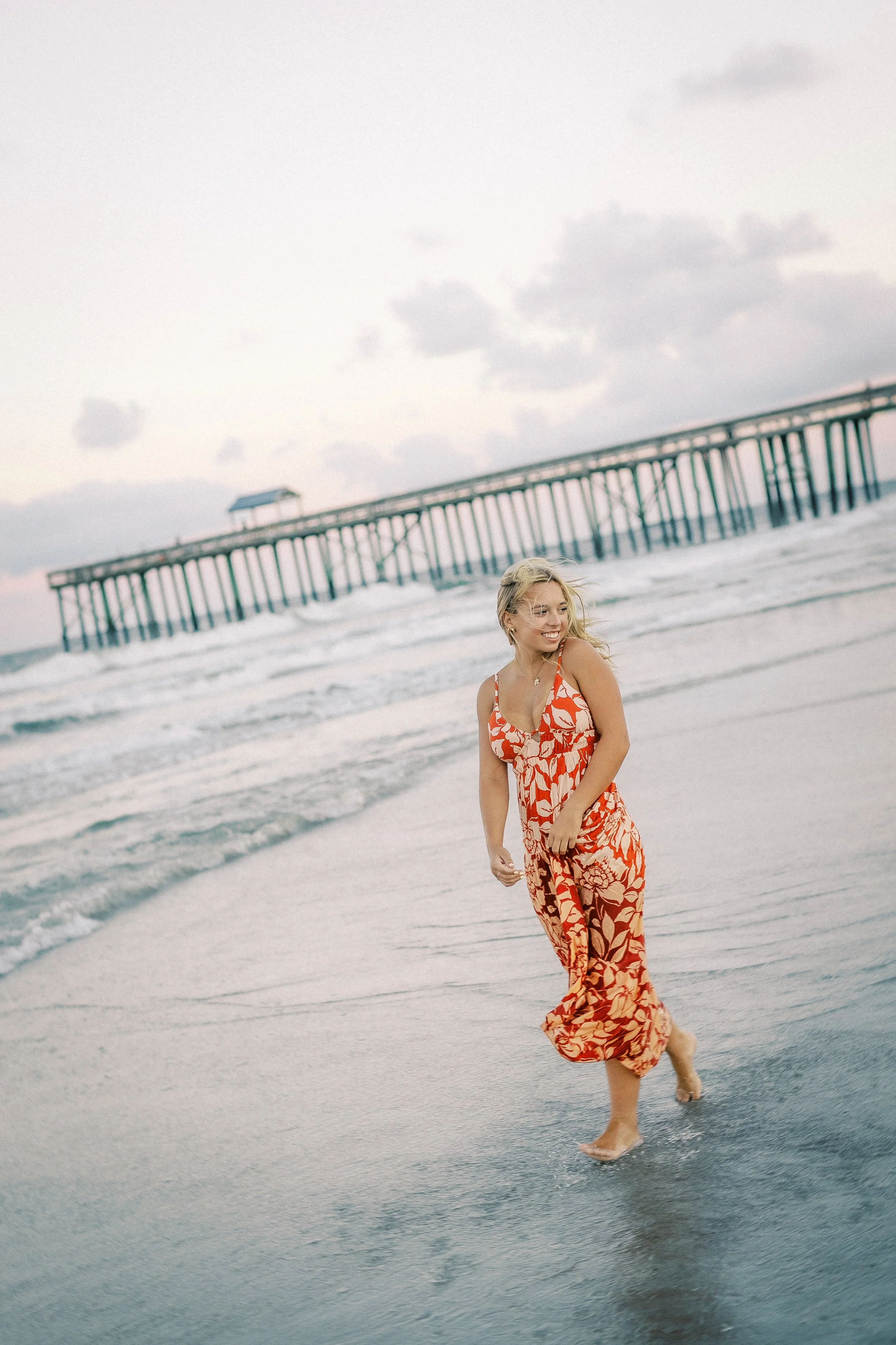 A woman in a red and orange floral dress walking barefoot along the wet beach shoreline, with a pier extending into the ocean in the background and cloudy sky above.