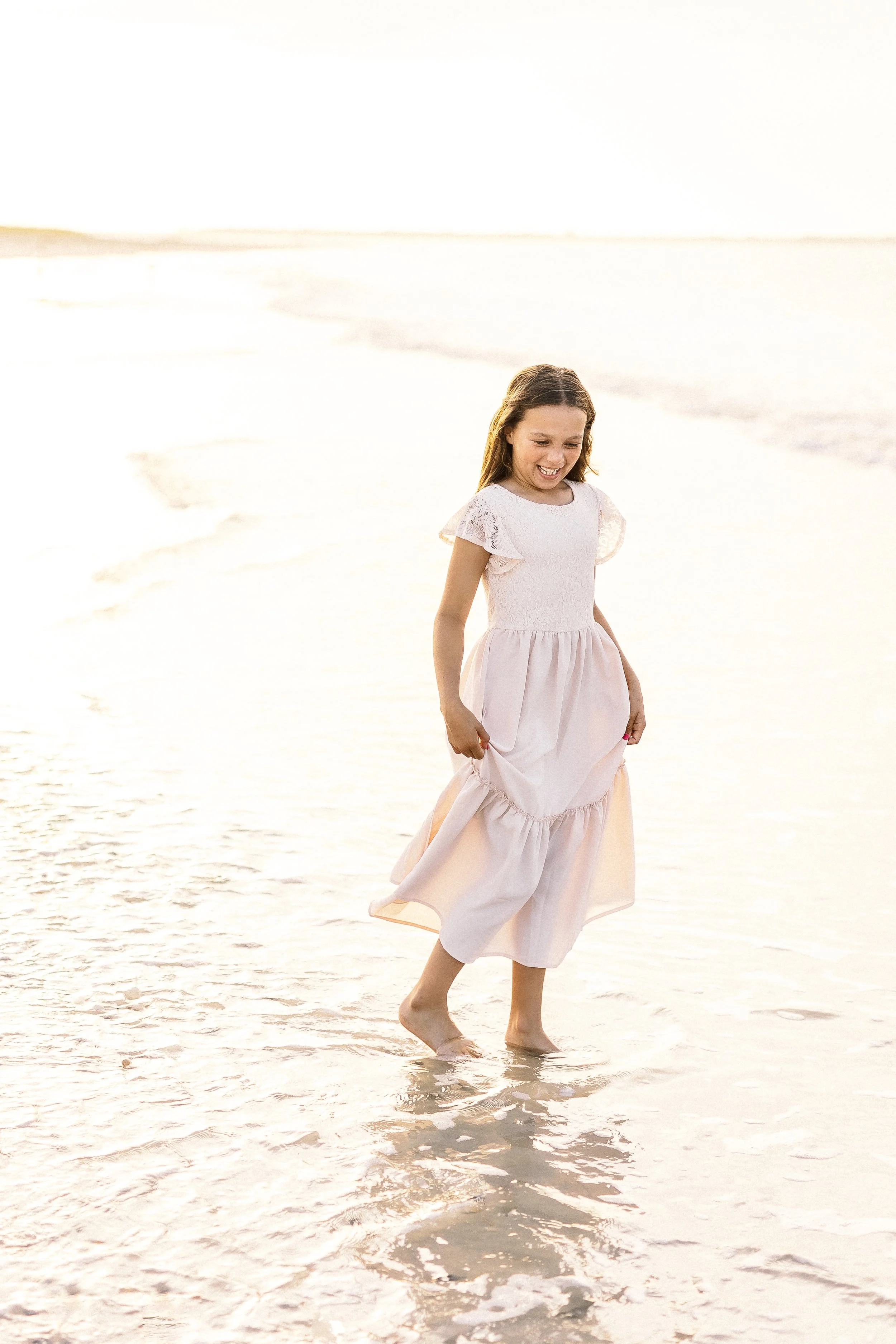 A young girl with long brown hair wearing a light pink dress and holding up her dress while walking barefoot in shallow ocean water at sunset.