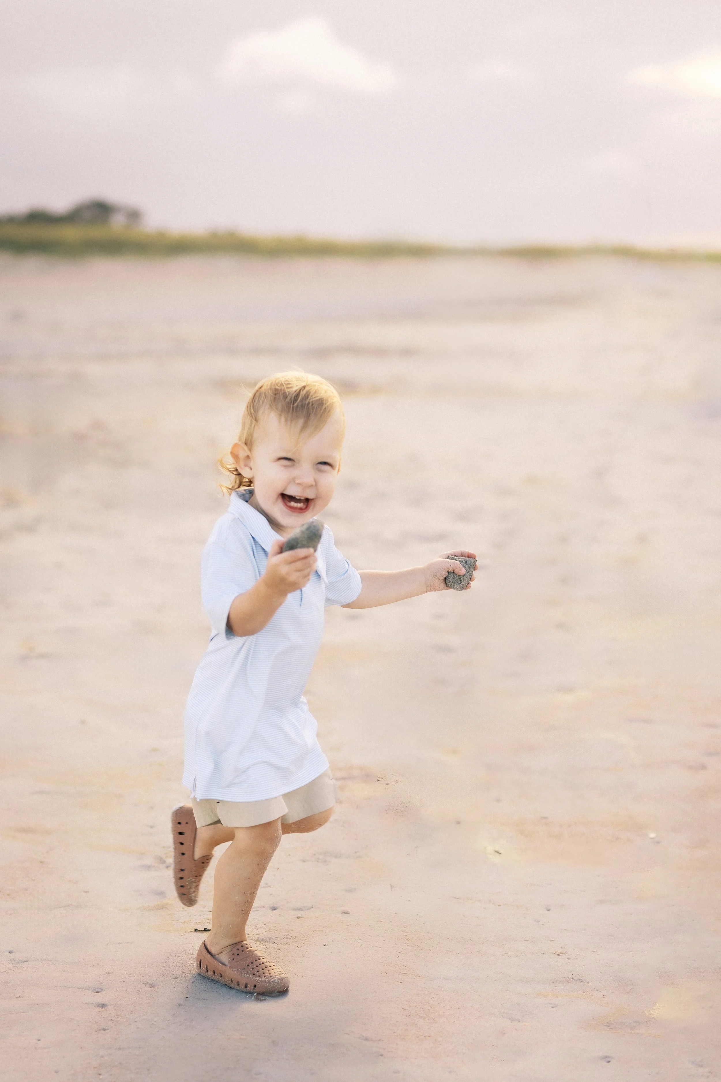 A young boy with blond hair and a big smile, running on the beach holding rocks in both hands, wearing a light striped shirt, beige shorts, and brown slip-on shoes. Photographer Amelia Island, Jacksonville, St Augustine, St Simons Island, Savannah