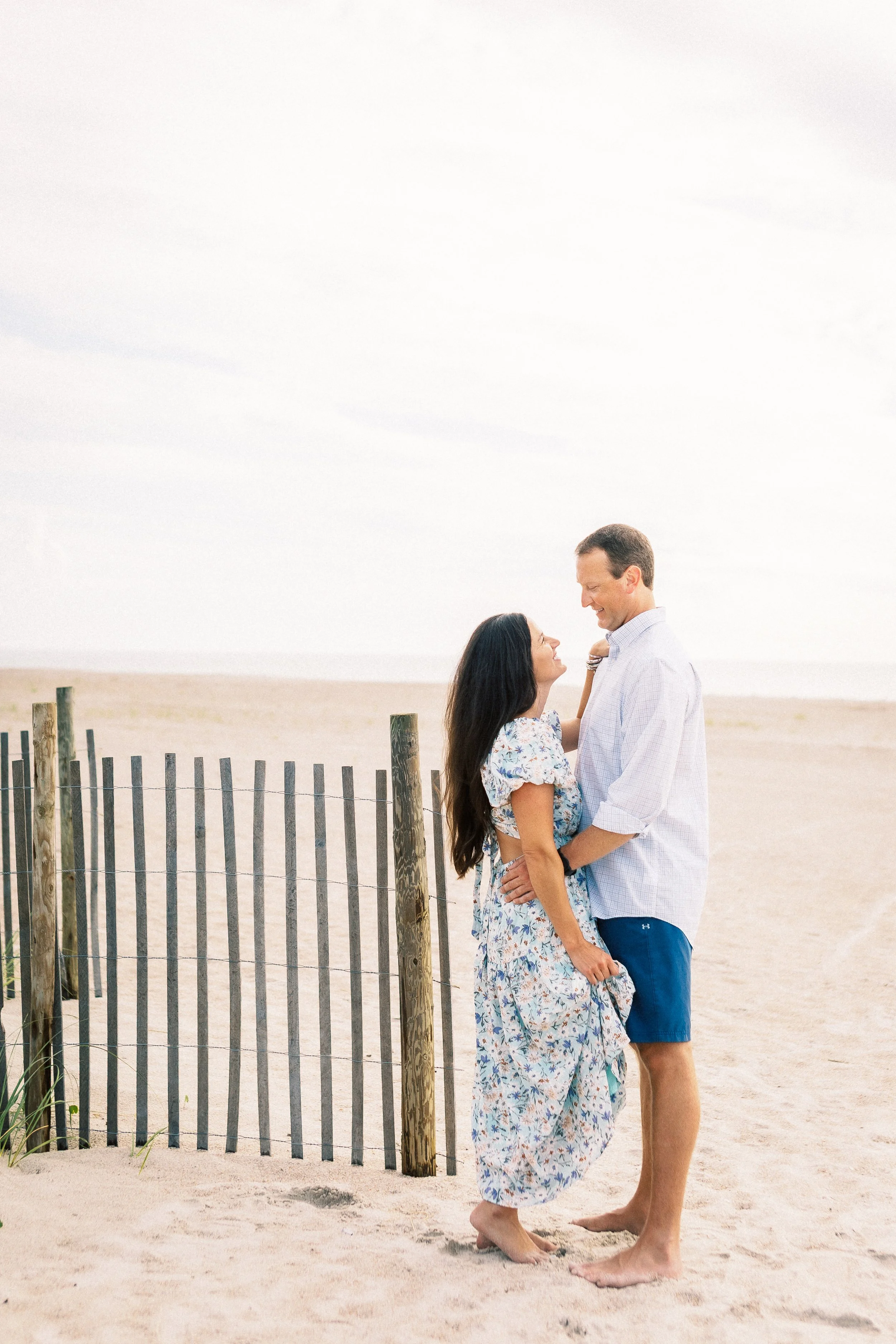 A couple standing on a sandy beach, facing each other and smiling, with a wooden fence behind them and a cloudy sky above.Photographer Amelia Island, Jacksonville, St Augustine, St Simons Island, Savannah photographer