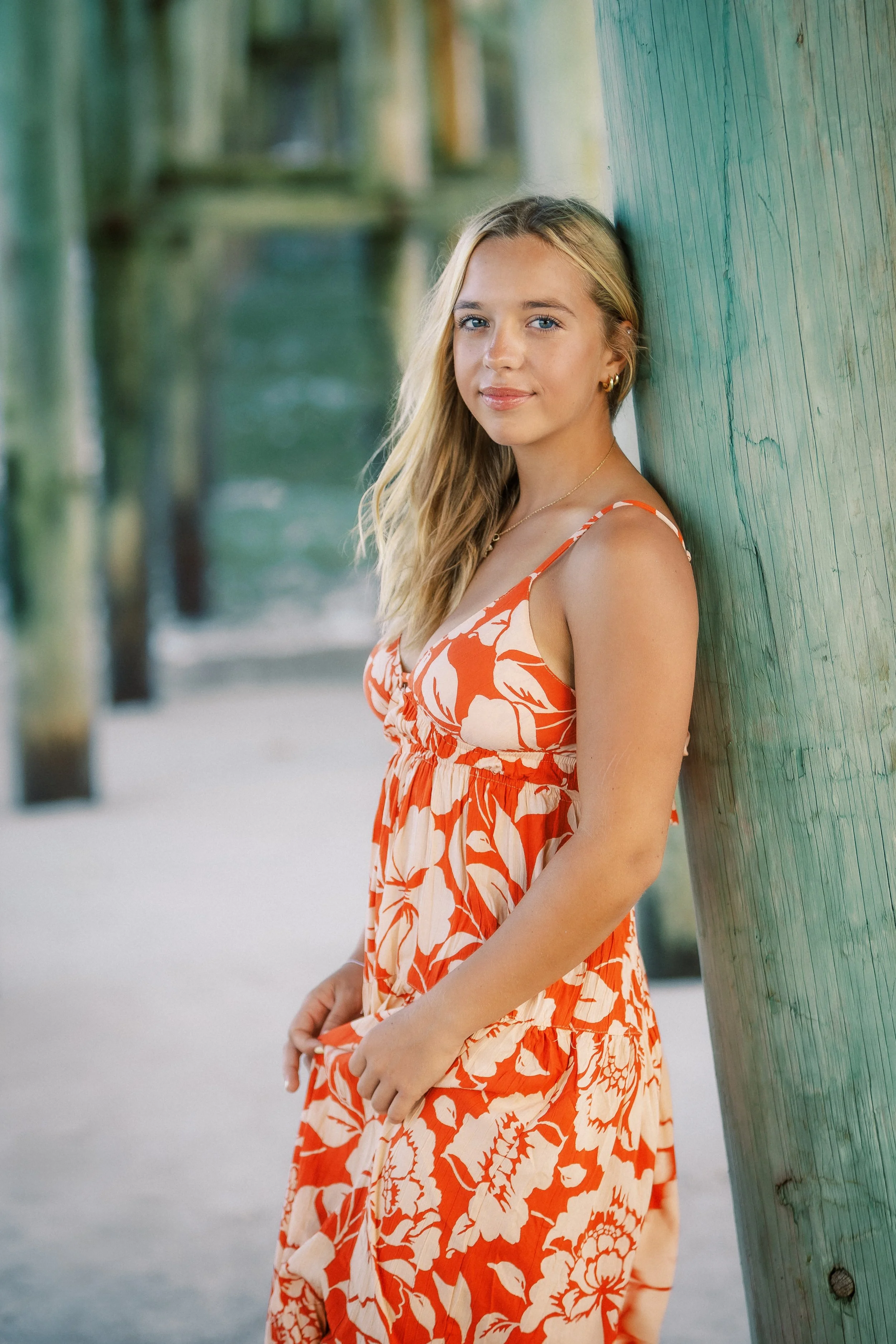 A young woman with long blonde hair, wearing an orange and white floral sundress, is standing and leaning against a teal wooden post on a beach. She has a slight smile and is looking at the camera, with a blurry background of more wooden posts and sa
