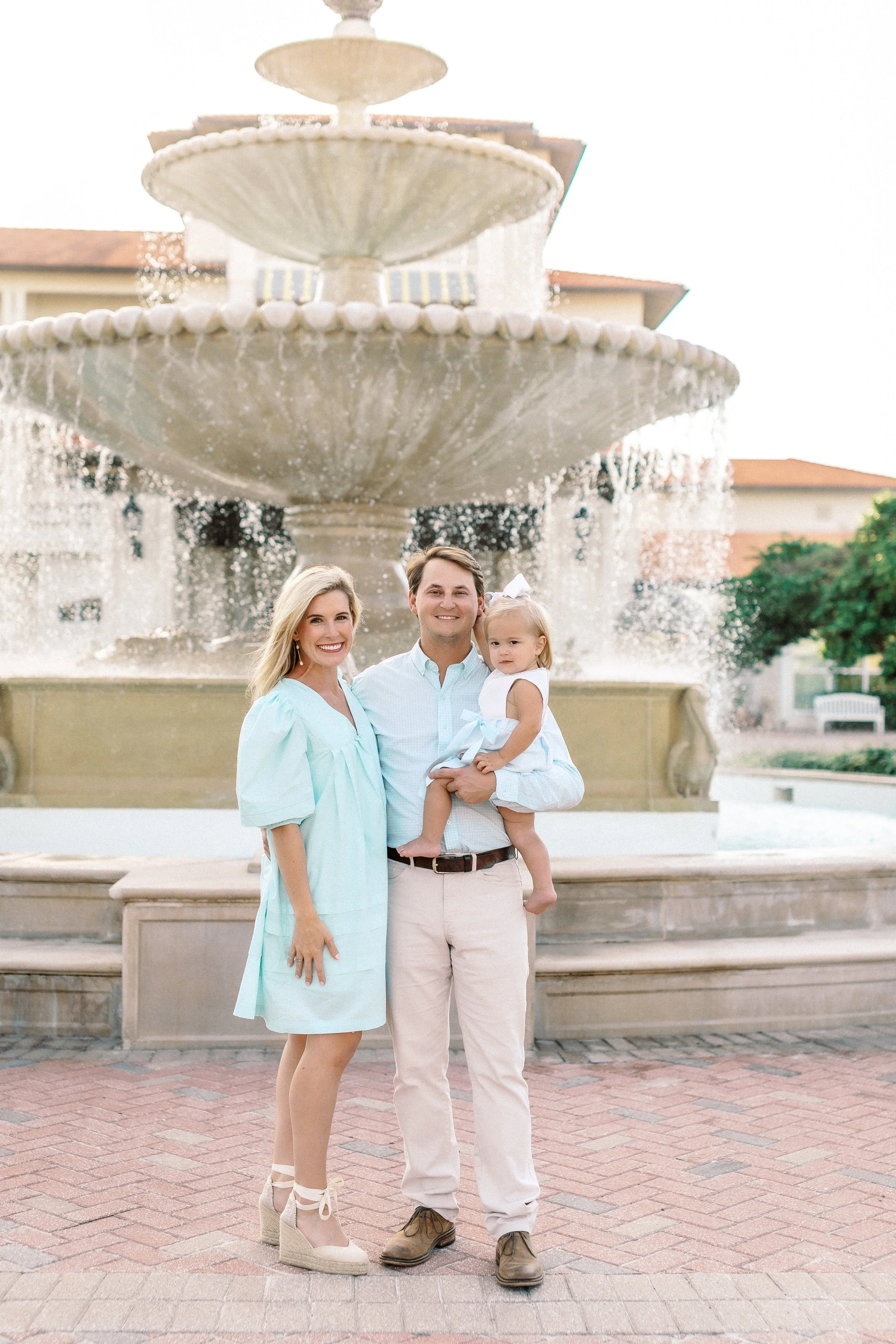 A family of three standing in front of a multi-tiered fountain, smiling. The woman is wearing a light blue dress with wedge heels, the man is in beige pants and brown shoes, and the young girl is in a white dress with a large white bow in her hair.