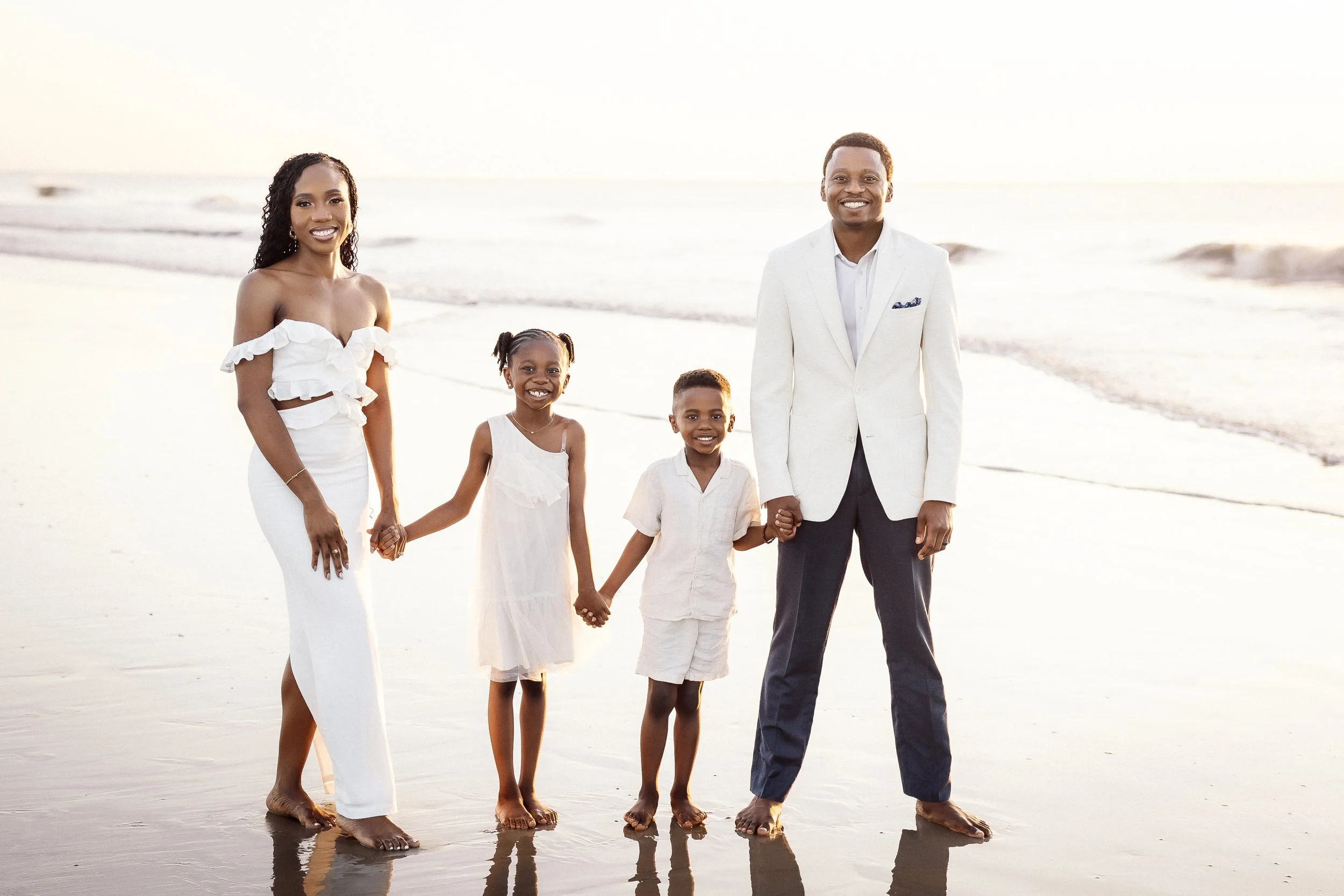 Family of five holding hands and walking on the beach during sunset, all dressed in white and smiling.