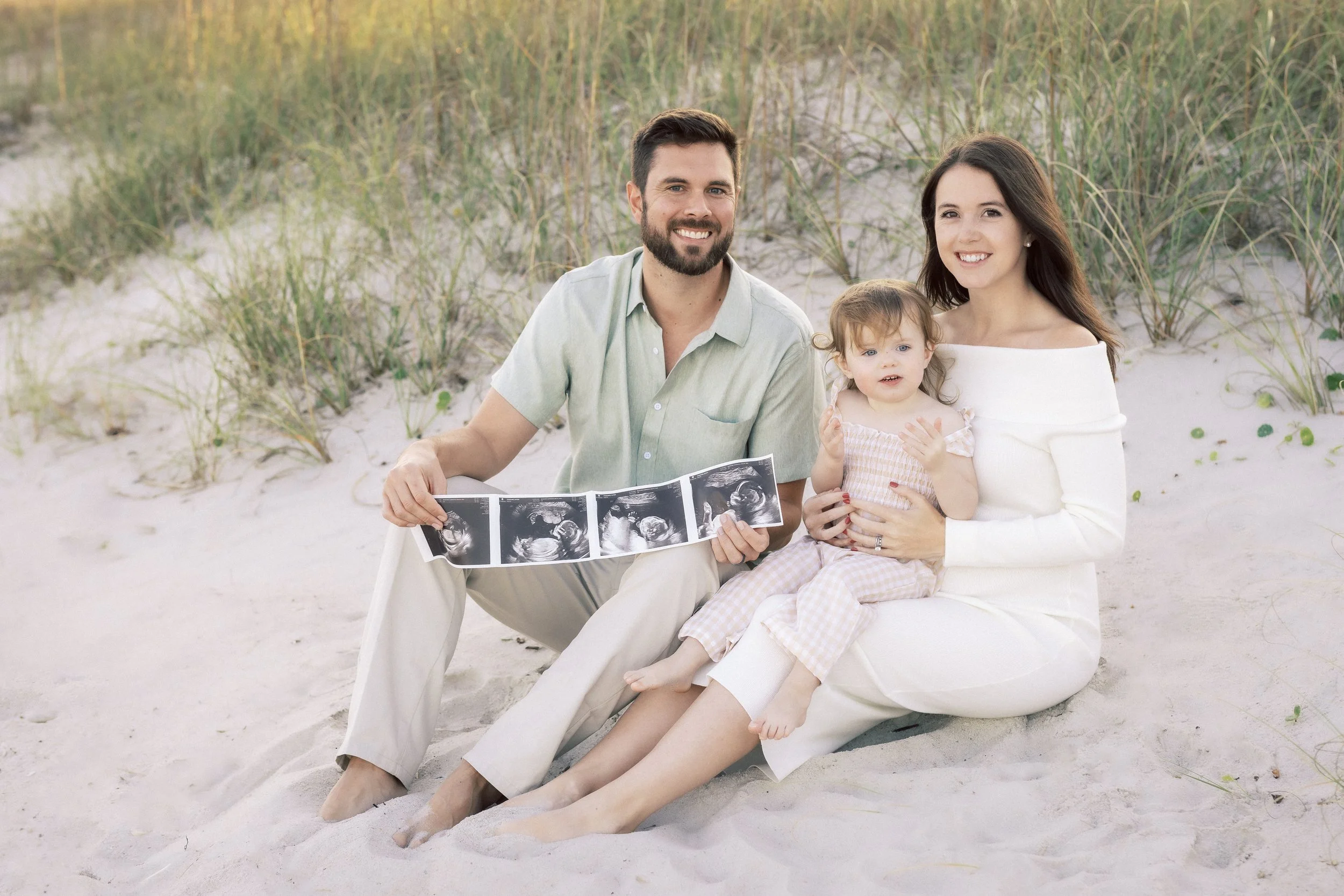 A family at the beach holding ultrasound images, with sand, grass, and dunes in the background.