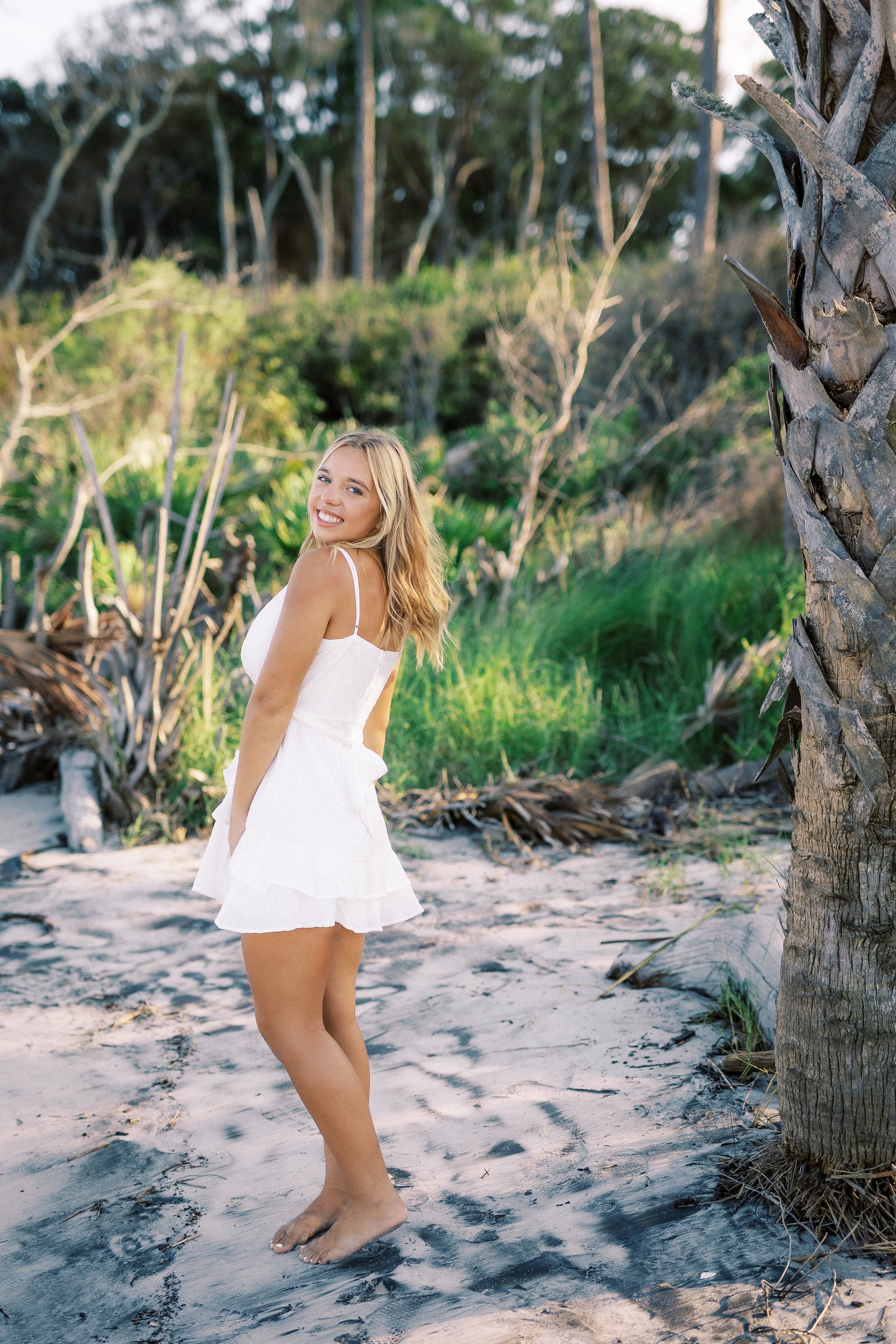 A young woman in a white dress standing barefoot on sandy beach with palm tree and lush green bushes in the background, smiling at the camera.