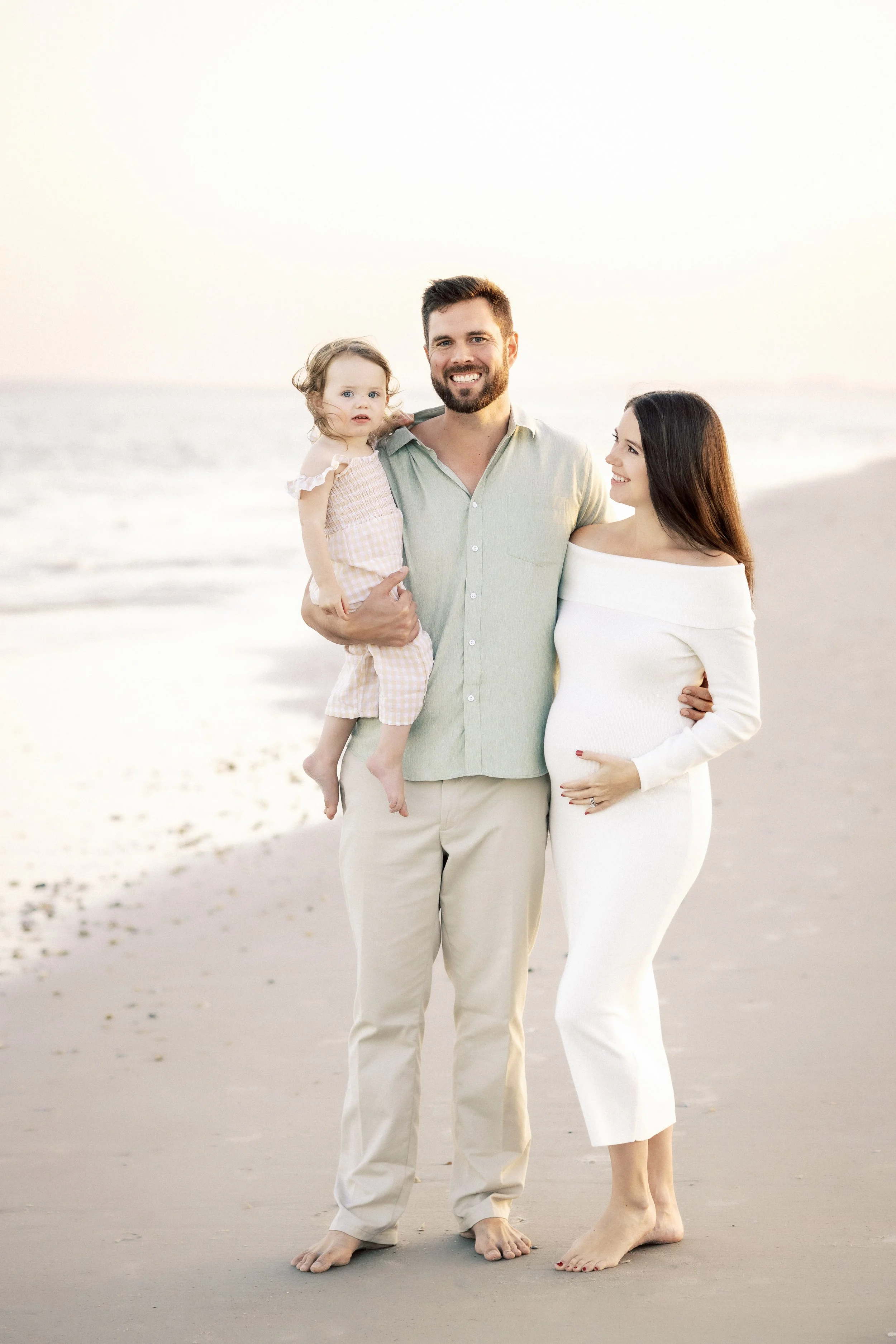A smiling man holding a young girl with curly hair, and a woman with long dark hair, standing barefoot on a sandy beach during sunset, with the ocean in the background.Photographer Amelia Island, Jacksonville, St Augustine, St Simons Island, Savannah
