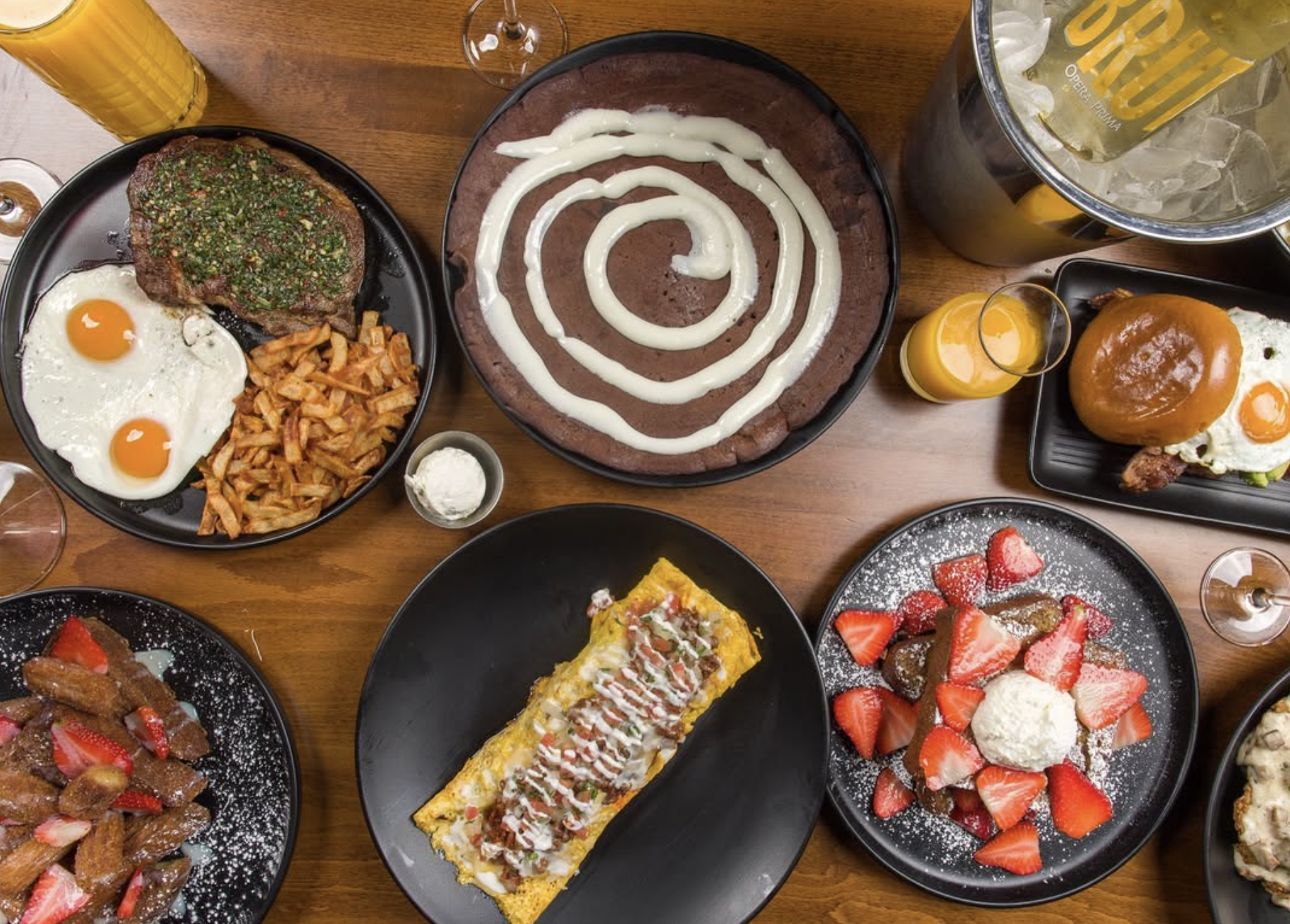 A variety of breakfast dishes on black plates including eggs, steak, hash browns, a pancake with white icing, a strawberry-topped dessert, a burrito, and a caramel syrup-topped dish, with glasses of orange juice and a bucket of cold drinks on a wooden table.