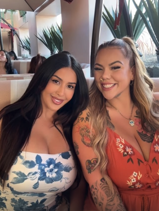 Two women sitting together at a restaurant or cafe, smiling and posing for the photo. The woman on the left has long dark hair, wearing a white dress with blue floral patterns. The woman on the right has long wavy blonde hair, wearing a red dress with floral embroidery and tattoos on her arms, with a septum piercing and jewelry.
